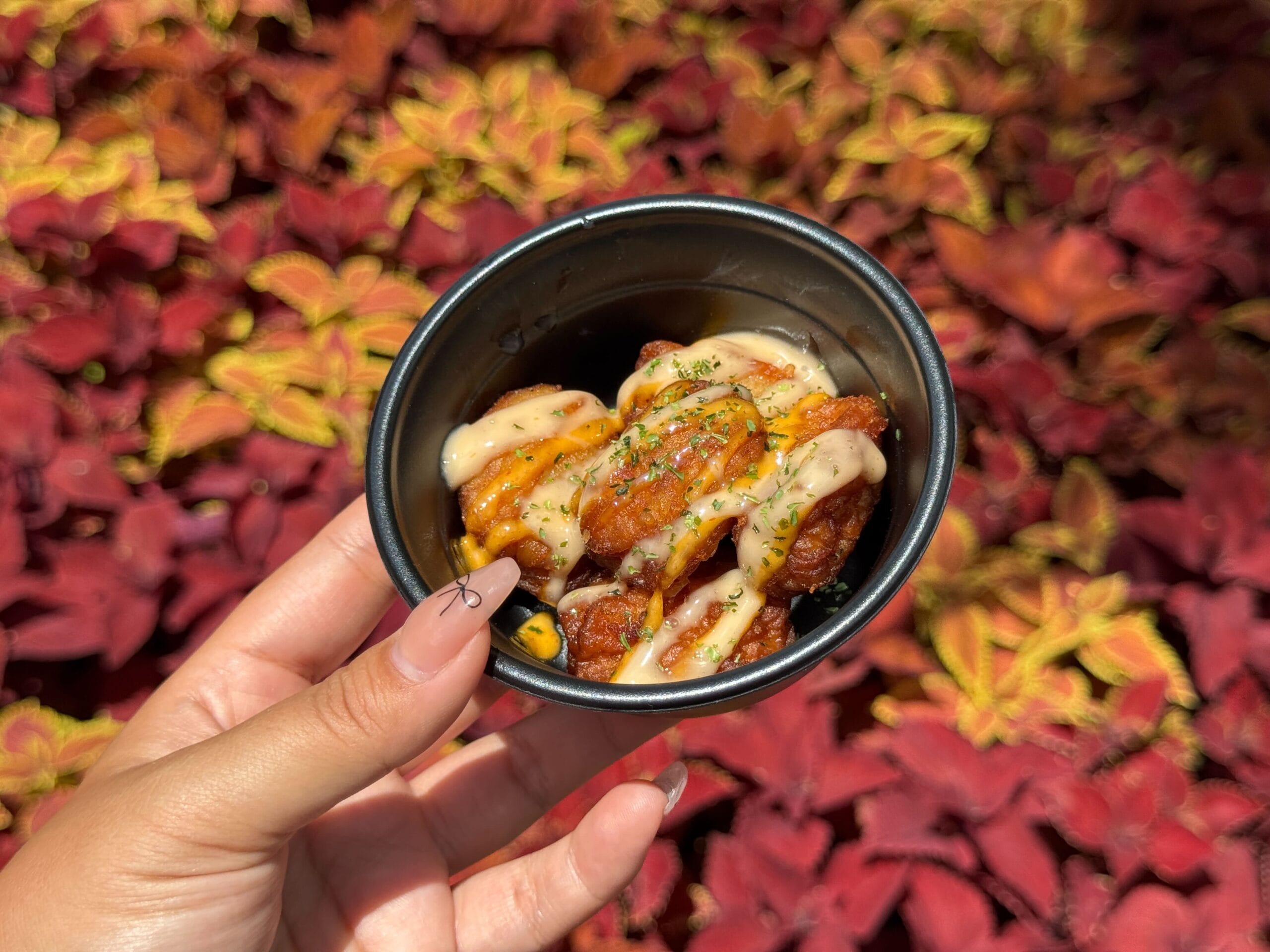 A hand holds a small bowl of sauced fried food, perfect for a Food & Wine moment against colorful foliage.
