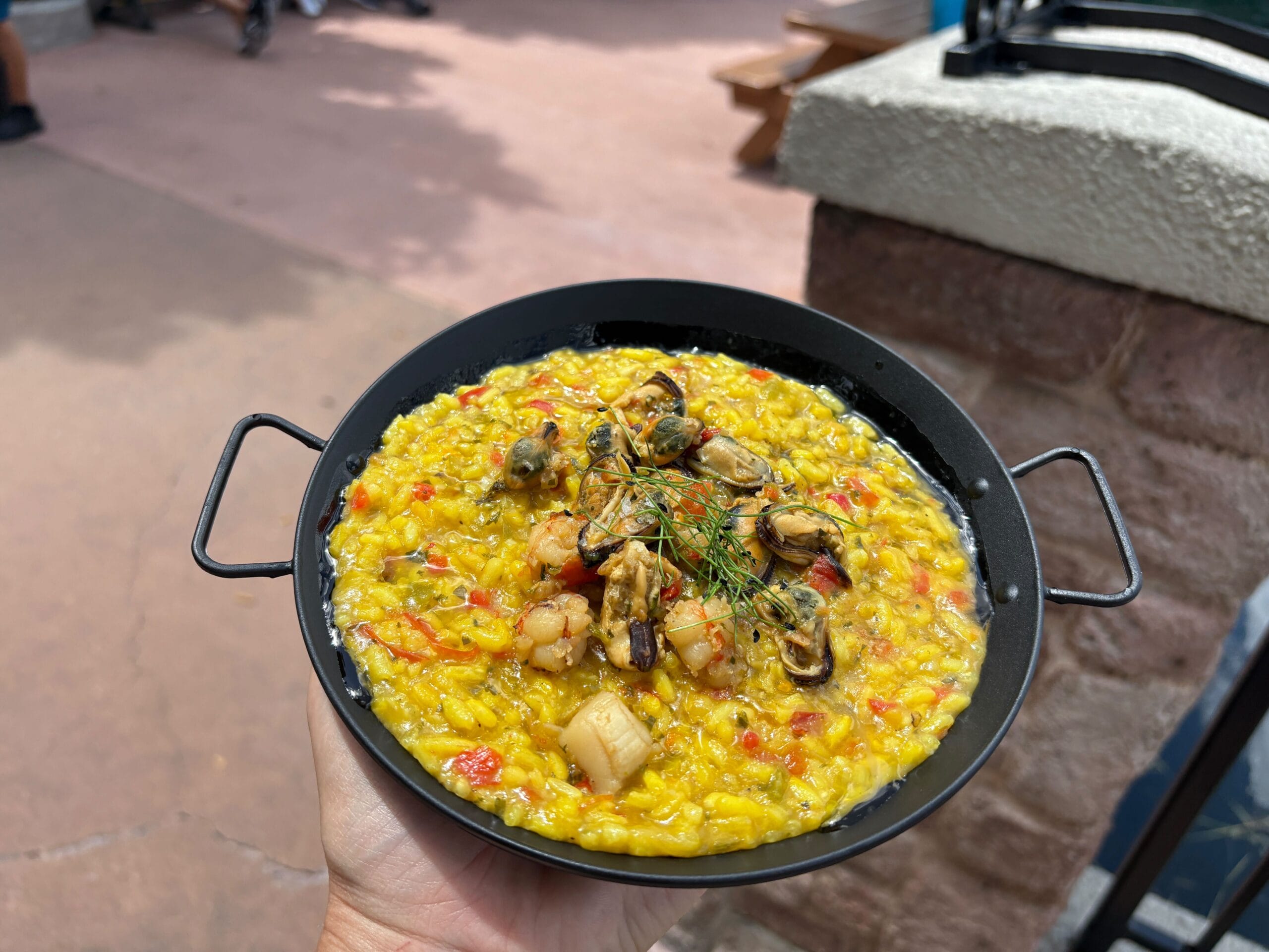 A hand holds a pan of yellow seafood risotto topped with shellfish and herbs, perfect for Food & Wine lovers, outdoors.