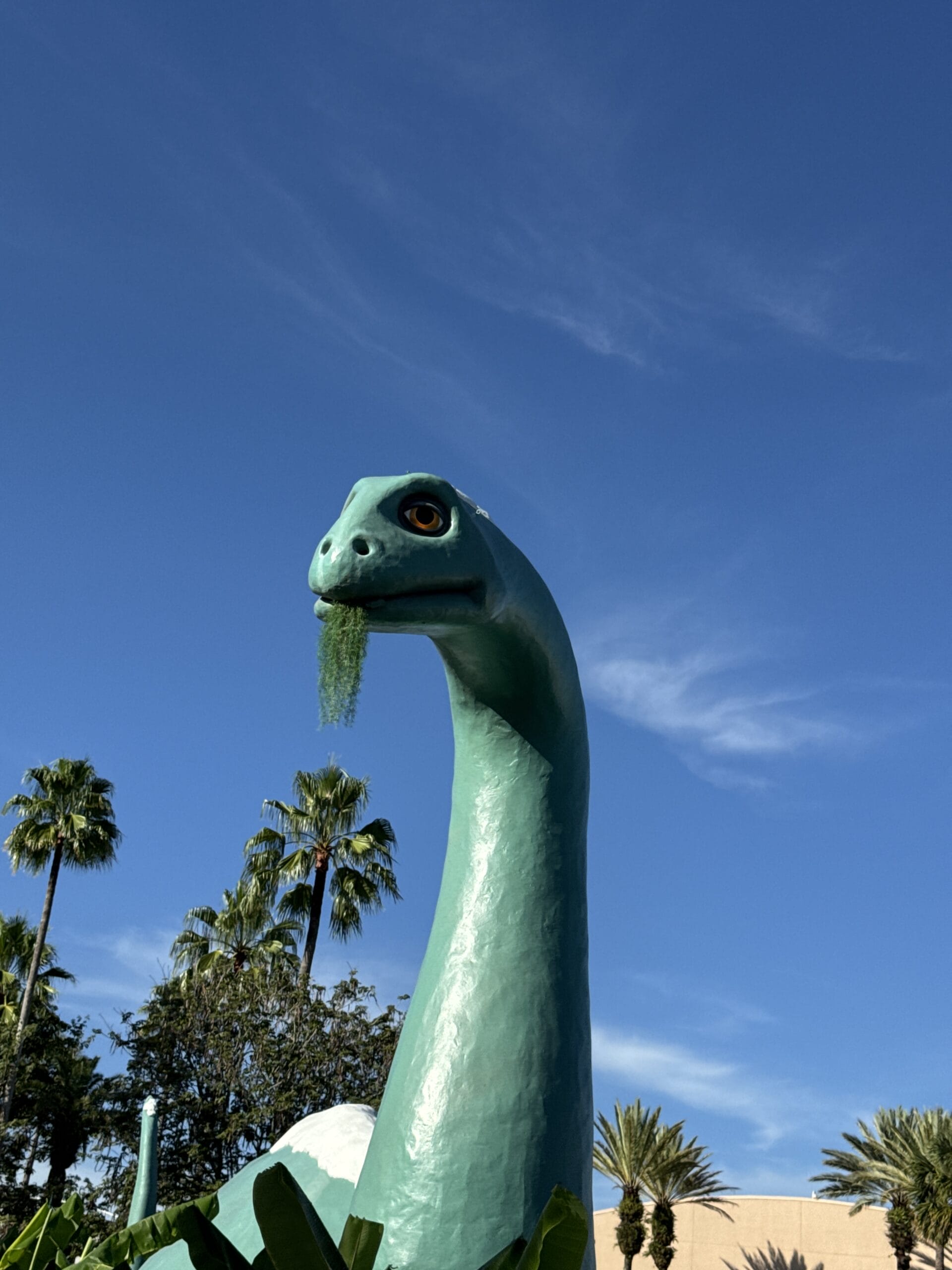 A large green dinosaur statue with a leafy mouth stands in Hollywood Studios among palm trees under a clear blue sky.