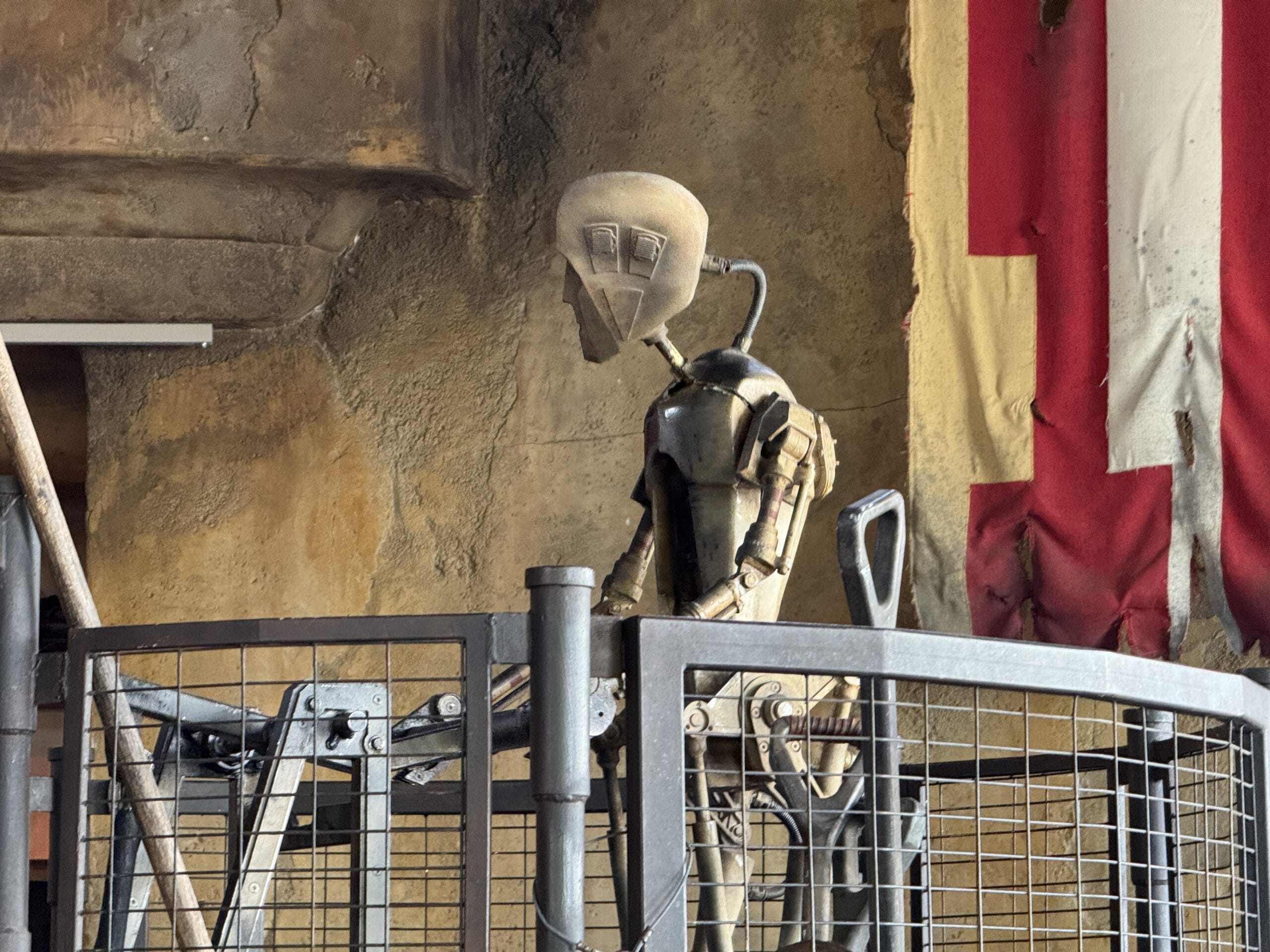 A humanoid robot stands behind a metal fence, as seen in a 2025 photo report from Hollywood Studios.