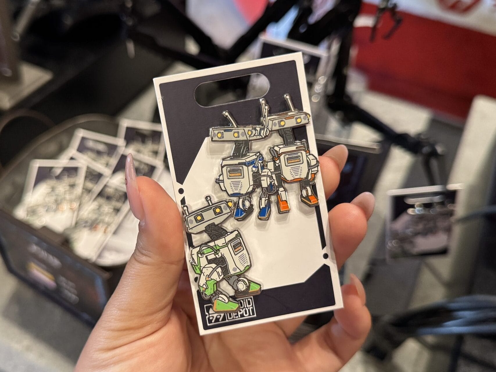 A hand holds a card with robot-themed enamel pins at Hollywood Studios, with more pins in the background.