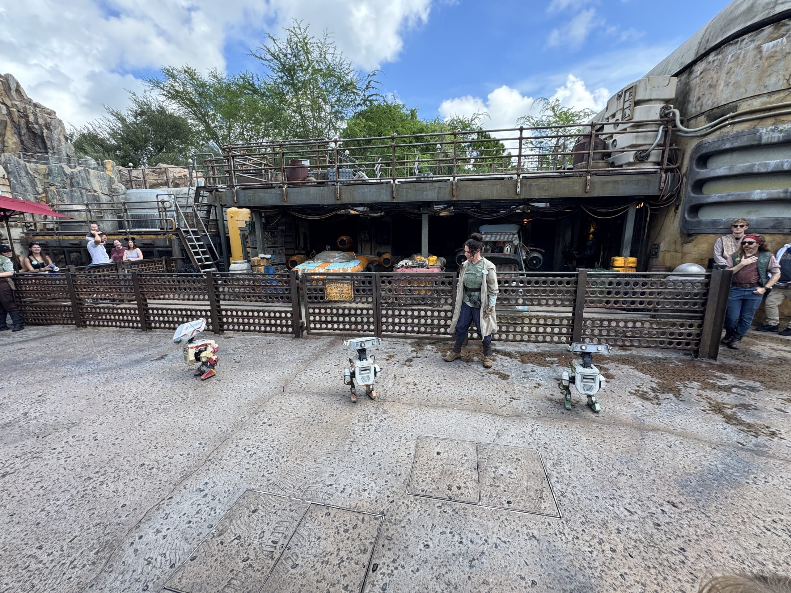 A person walks in front of a sci-fi themed building at Hollywood Studios, with three small robot figures in this 2025 photo report.