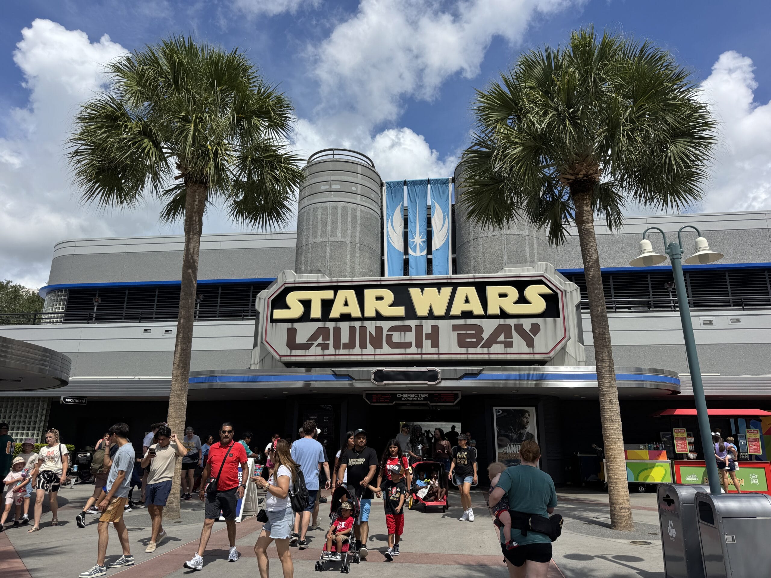 Guests stroll past the Star Wars Launch Bay at Hollywood Studios under palm trees and a bright blue 2025 sky.