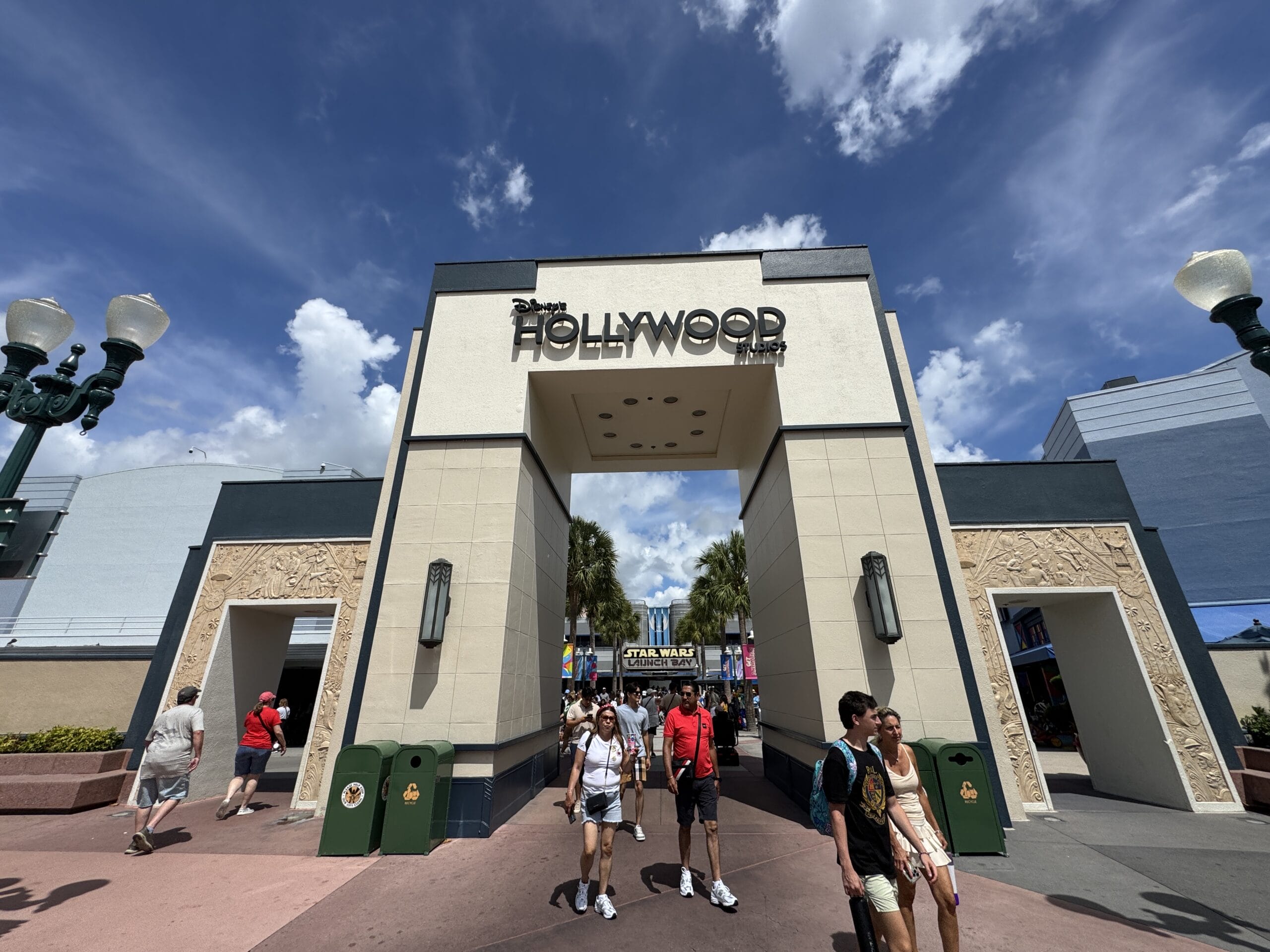 Guests walk under the “Hollywood” arch at Hollywood Studios on a sunny, partly cloudy day in this 2025 photo report.