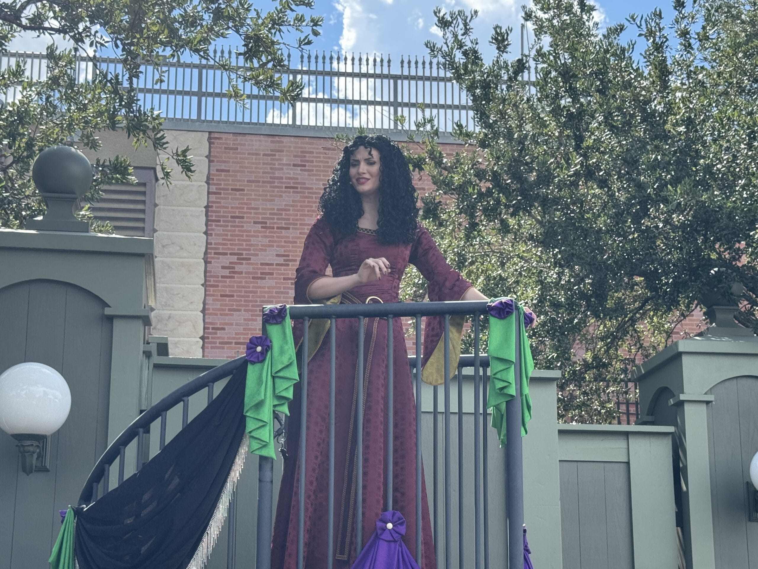 A woman in a maroon dress stands on a decorated outdoor staircase at Oogie Boogie Bash, surrounded by greenery and brick walls.