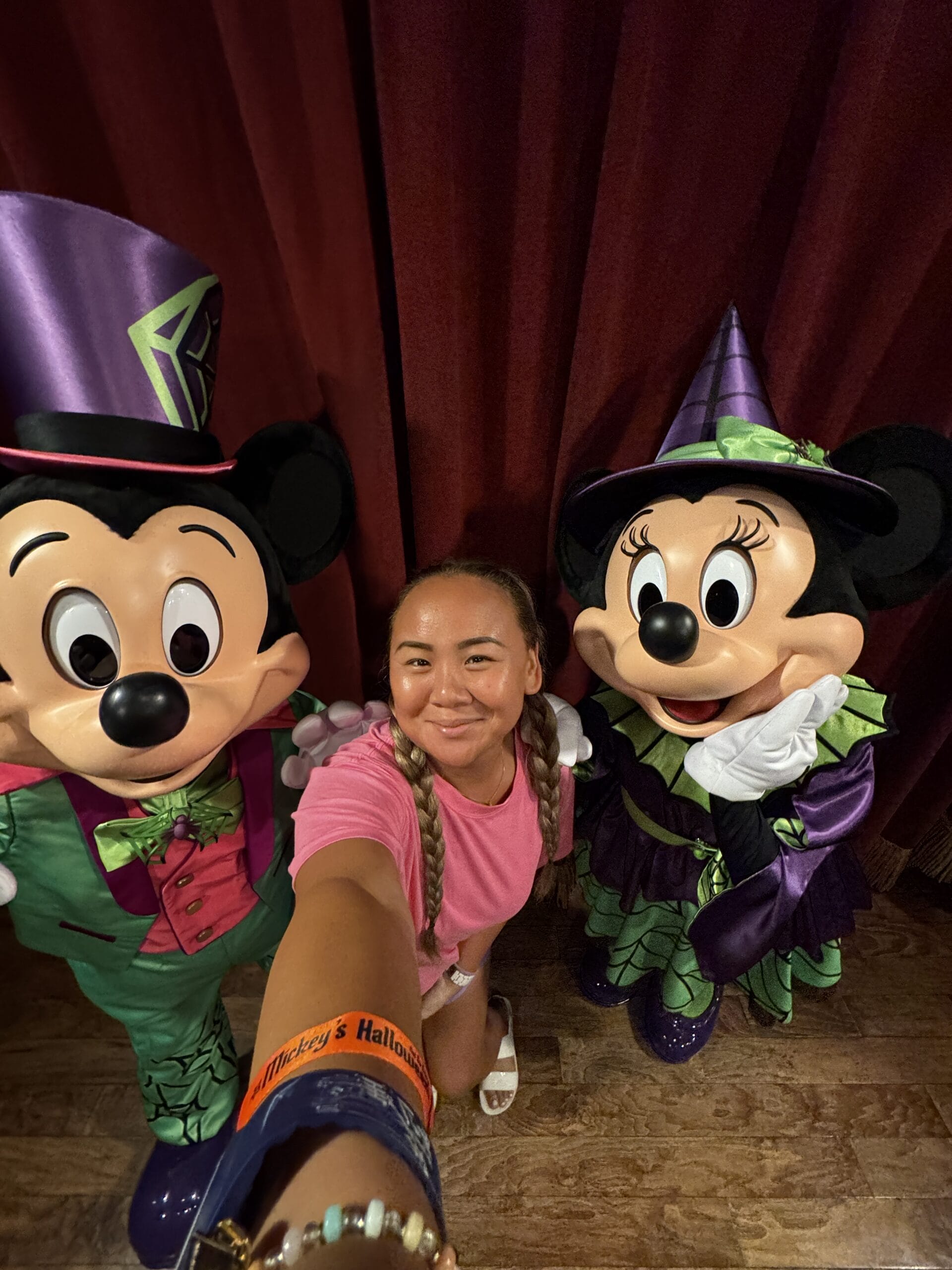 A woman takes a selfie with Mickey and Minnie Mouse at Oogie Boogie Bash in front of red curtains.