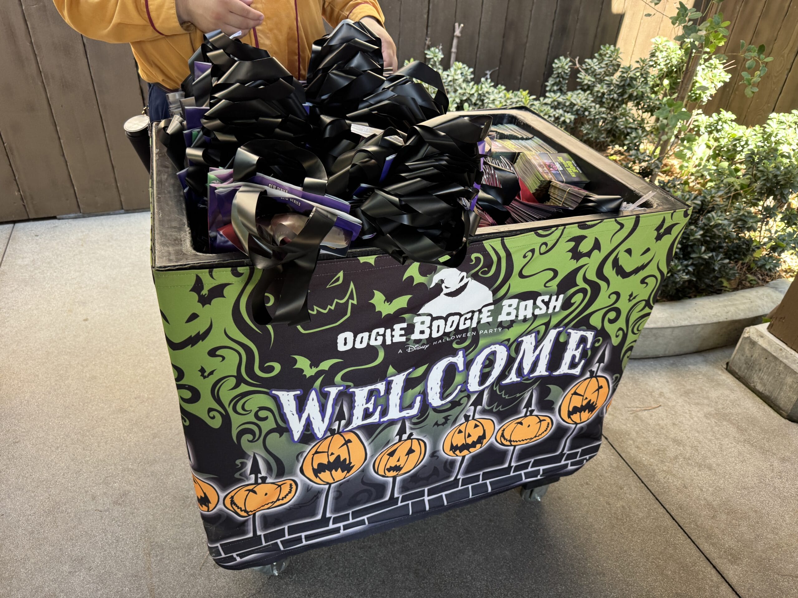 A photo report of a Halloween cart with "Oogie Boogie Bash Welcome" and bags handed out by a person in a yellow jacket.