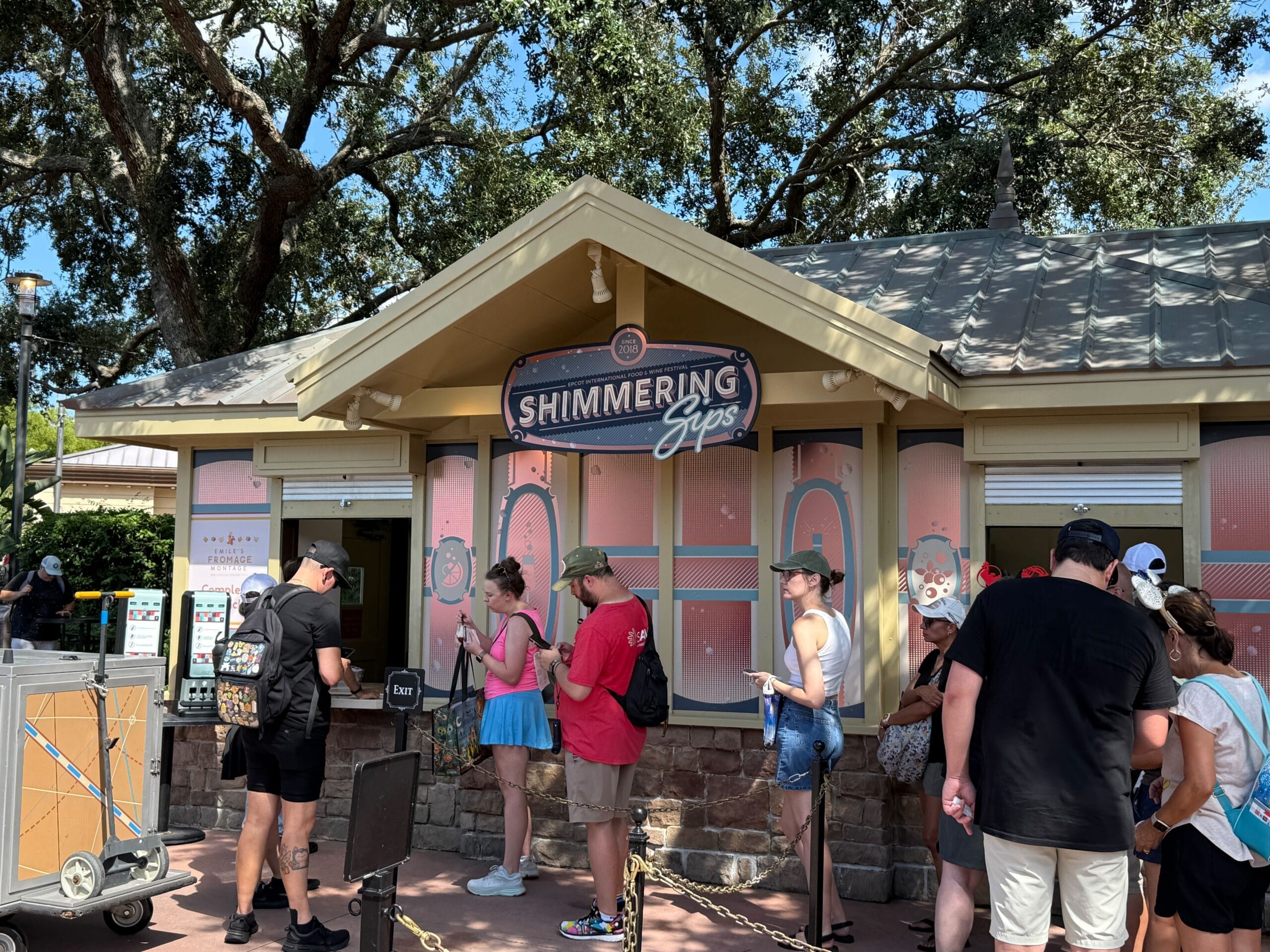 People wait in line at the Shimmering Sips food booth, eager to try New Food & Wine under sunny trees.
