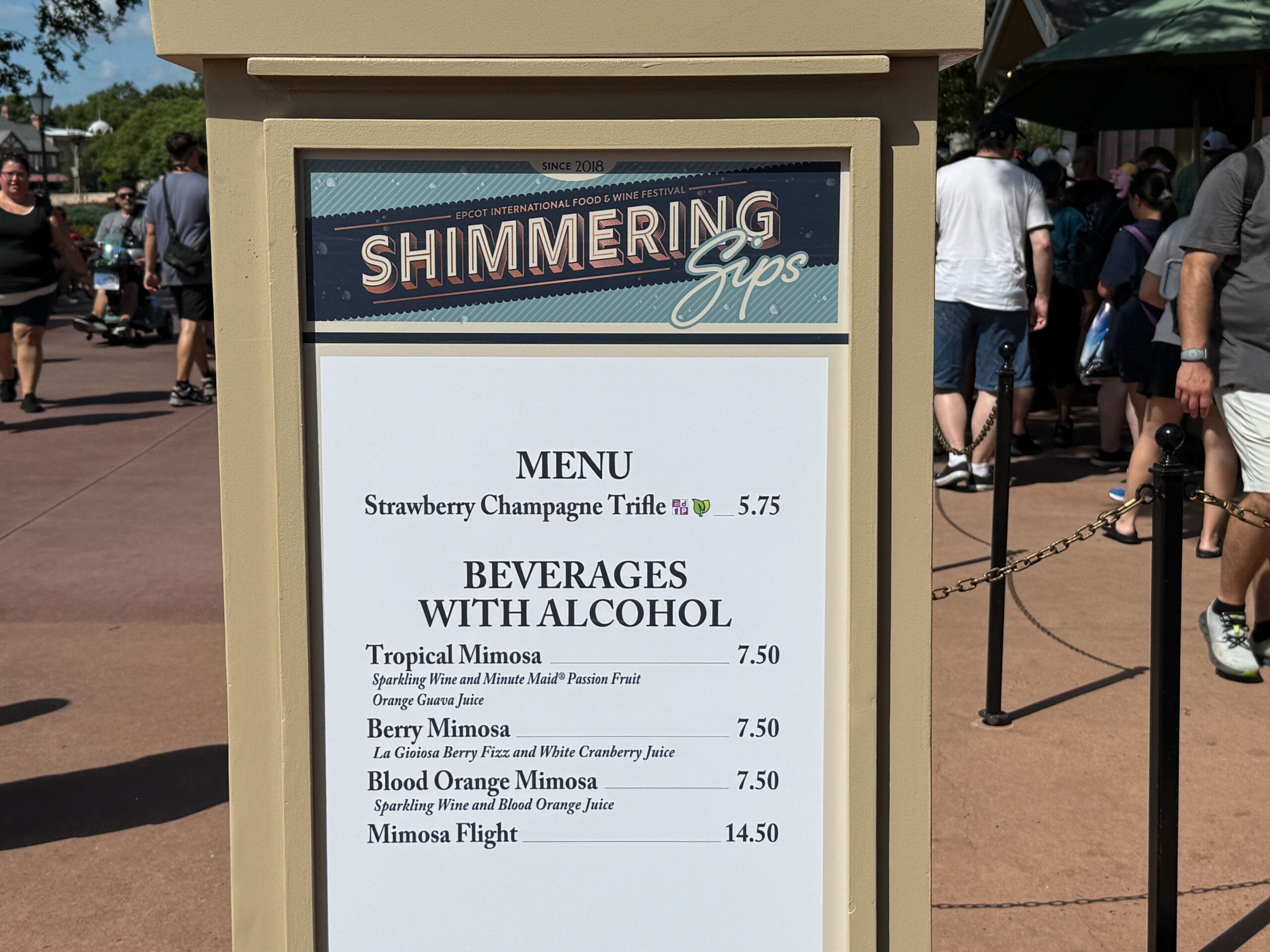Outdoor menu board for "Shimmering Sips" highlights New Food & Wine desserts and drinks, with people in the background.