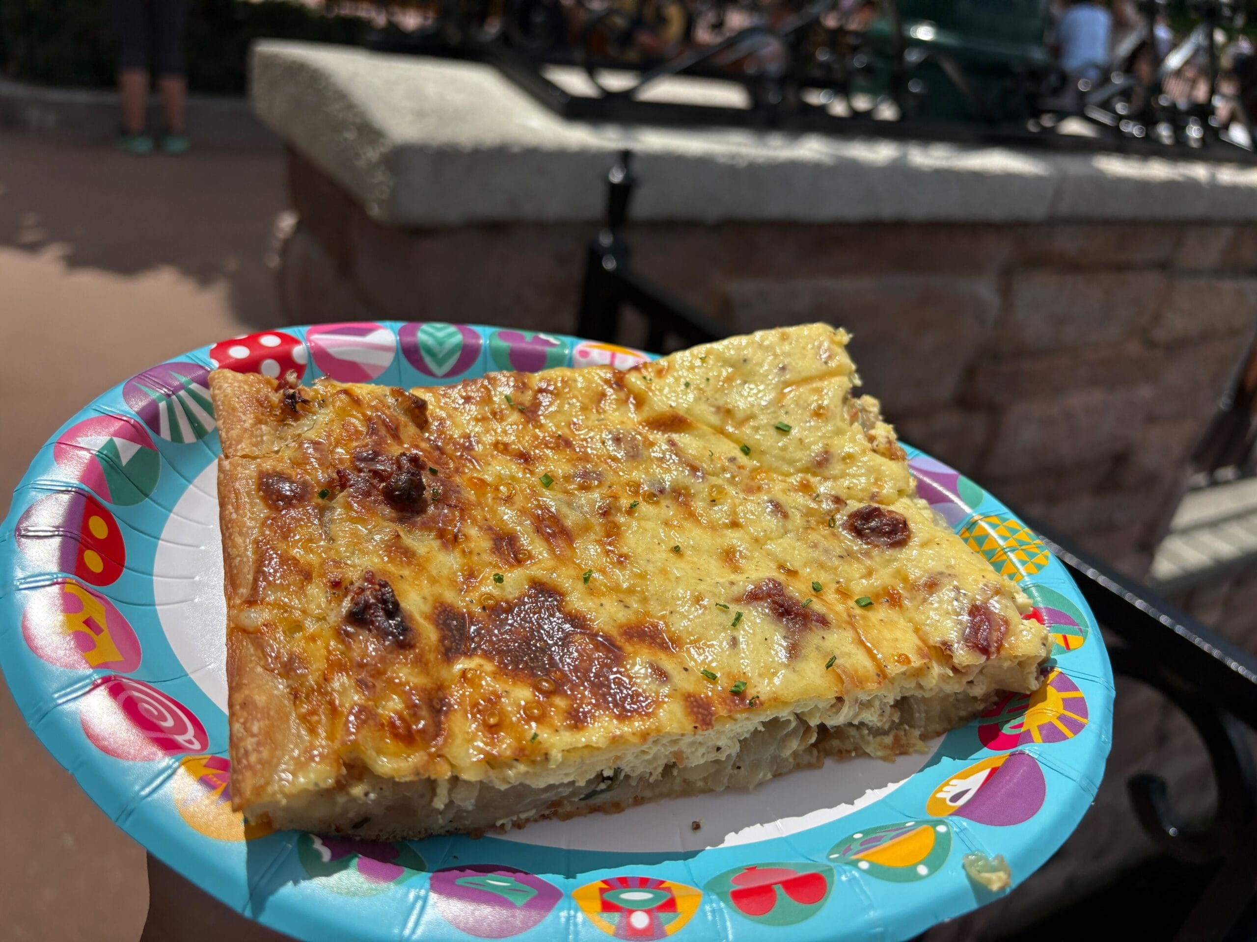 A hand holds a colorful paper plate with a large slice of cheesy, rectangular pizza outdoors at a Food & Wine event.