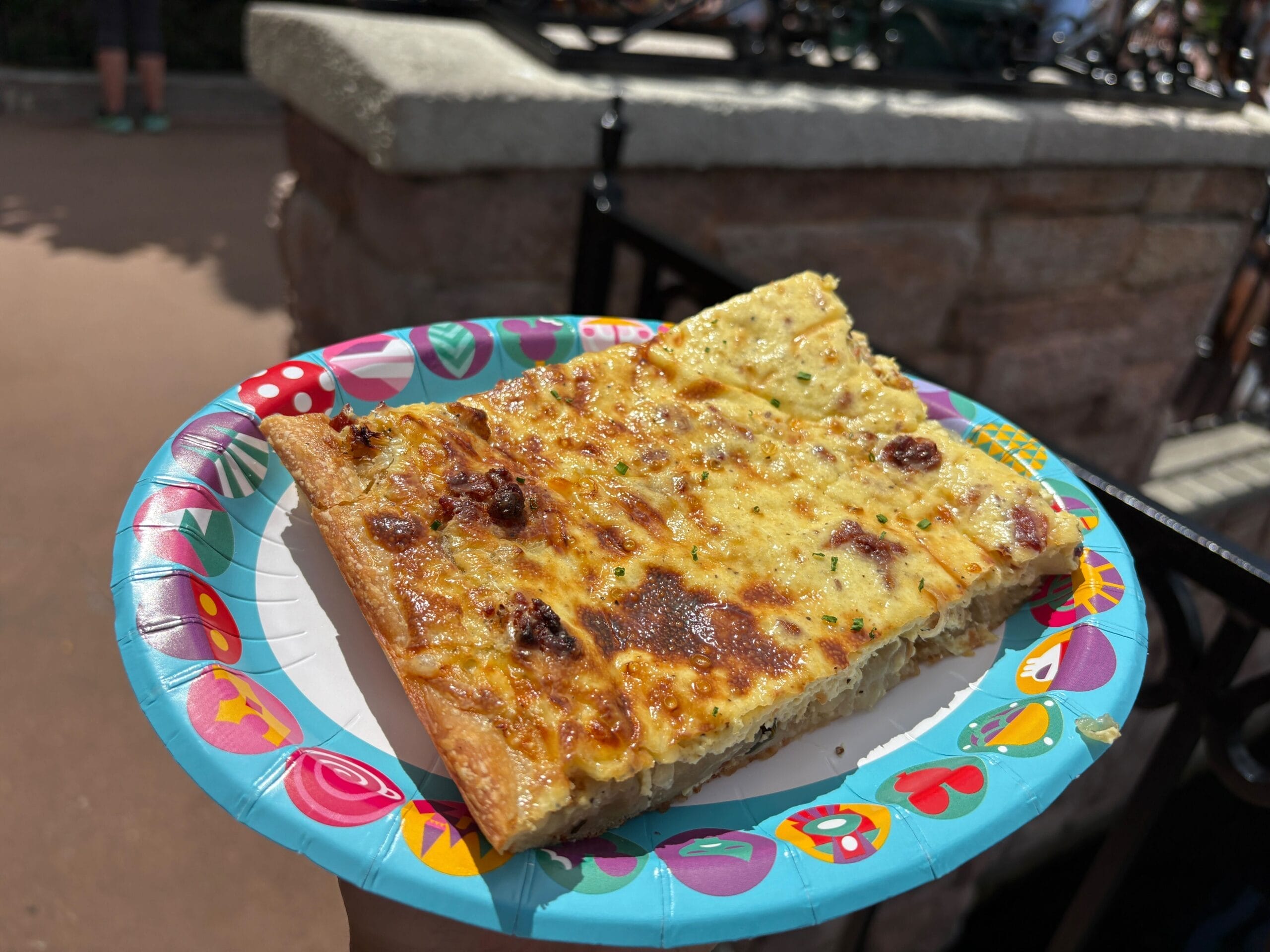 A slice of cheesy flatbread pizza on a colorful plate at the 2025 EPCOT Food and Wine Festival, outdoors by a stone ledge.