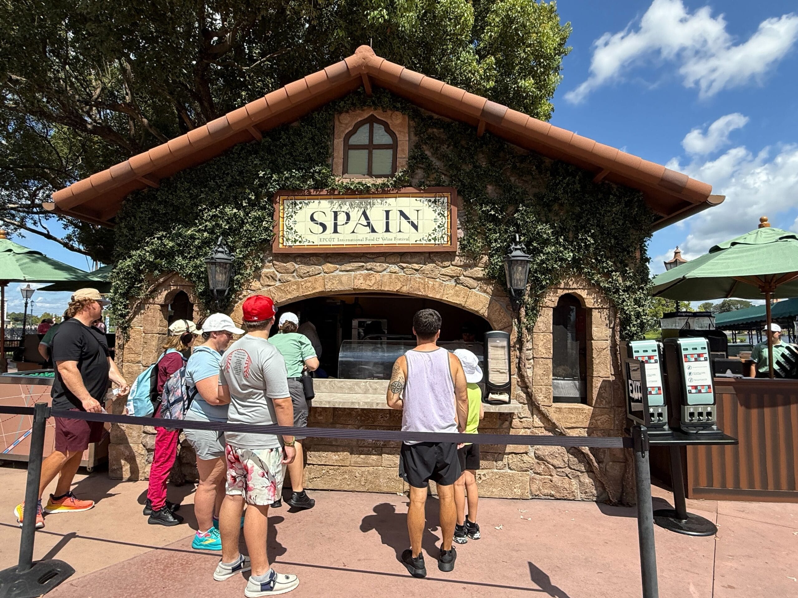 People line up at a Spain-themed Food & Wine booth with a stone exterior and a "SPAIN" sign above the window.