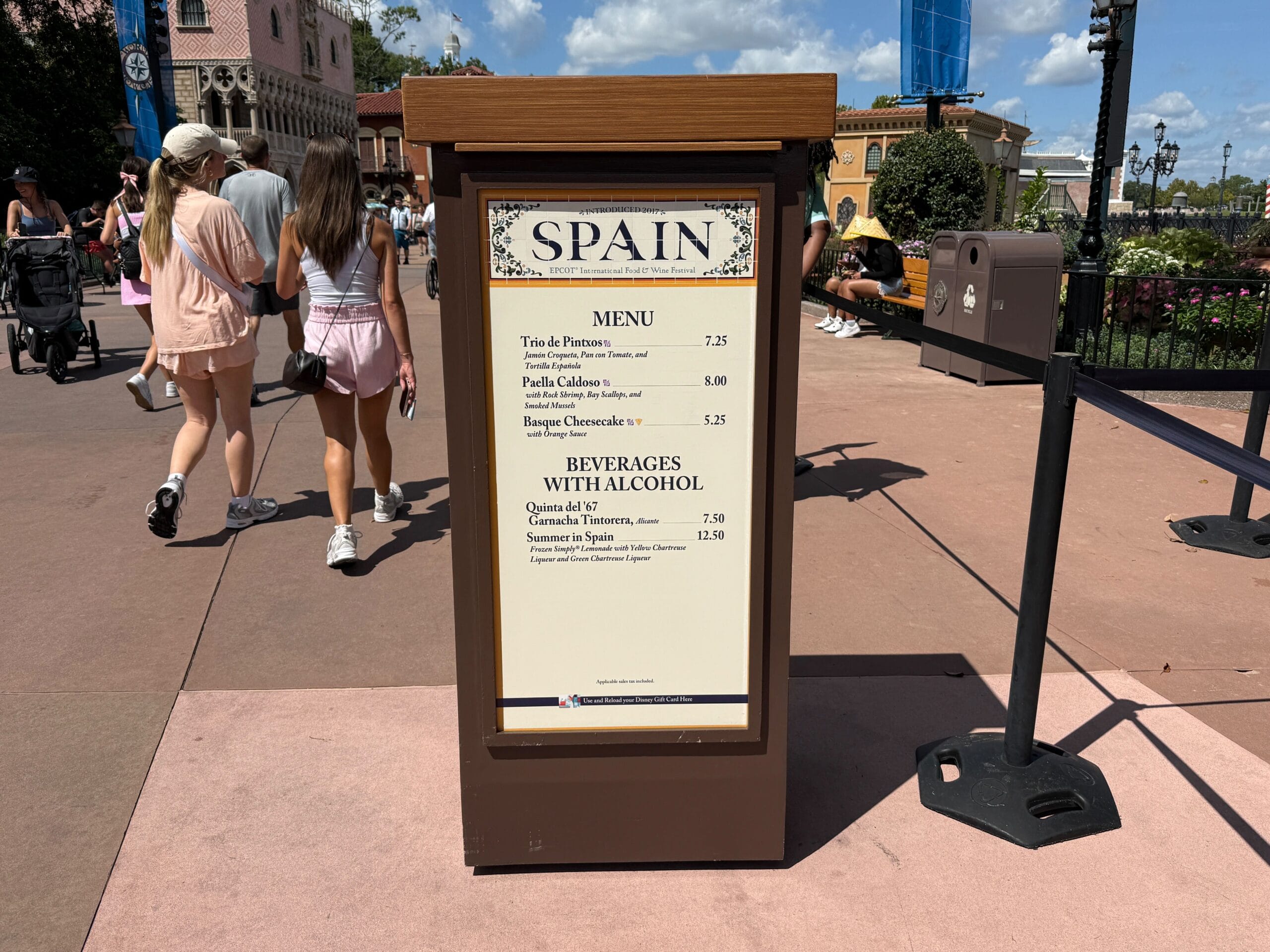 A Spain Food & Wine booth menu sign with people walking by at an outdoor event under a sunny sky.
