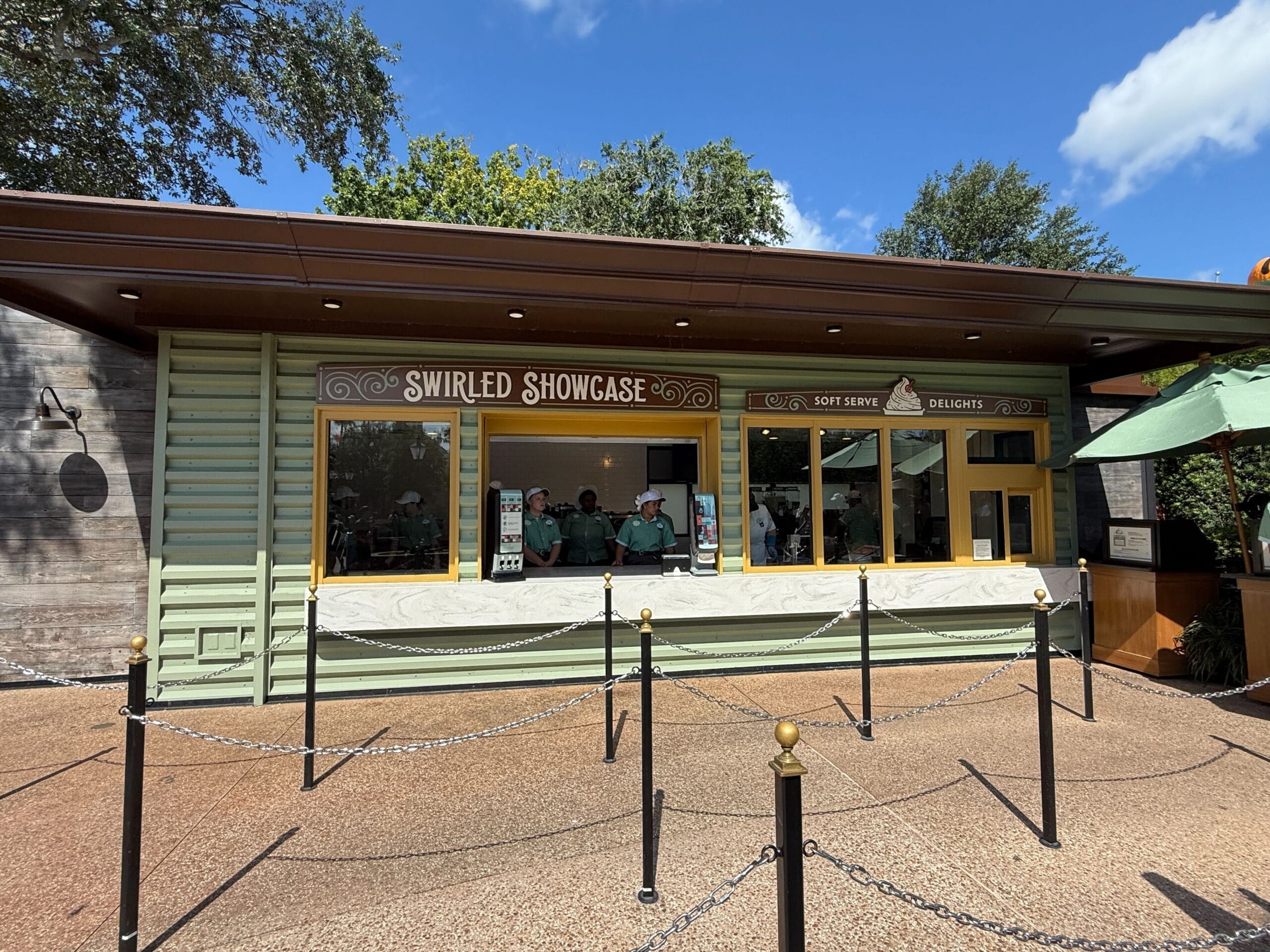 Swirled Showcase stand with menu boards and staff inside, plus New Food & Wine, outdoor queue under sunny sky and trees.