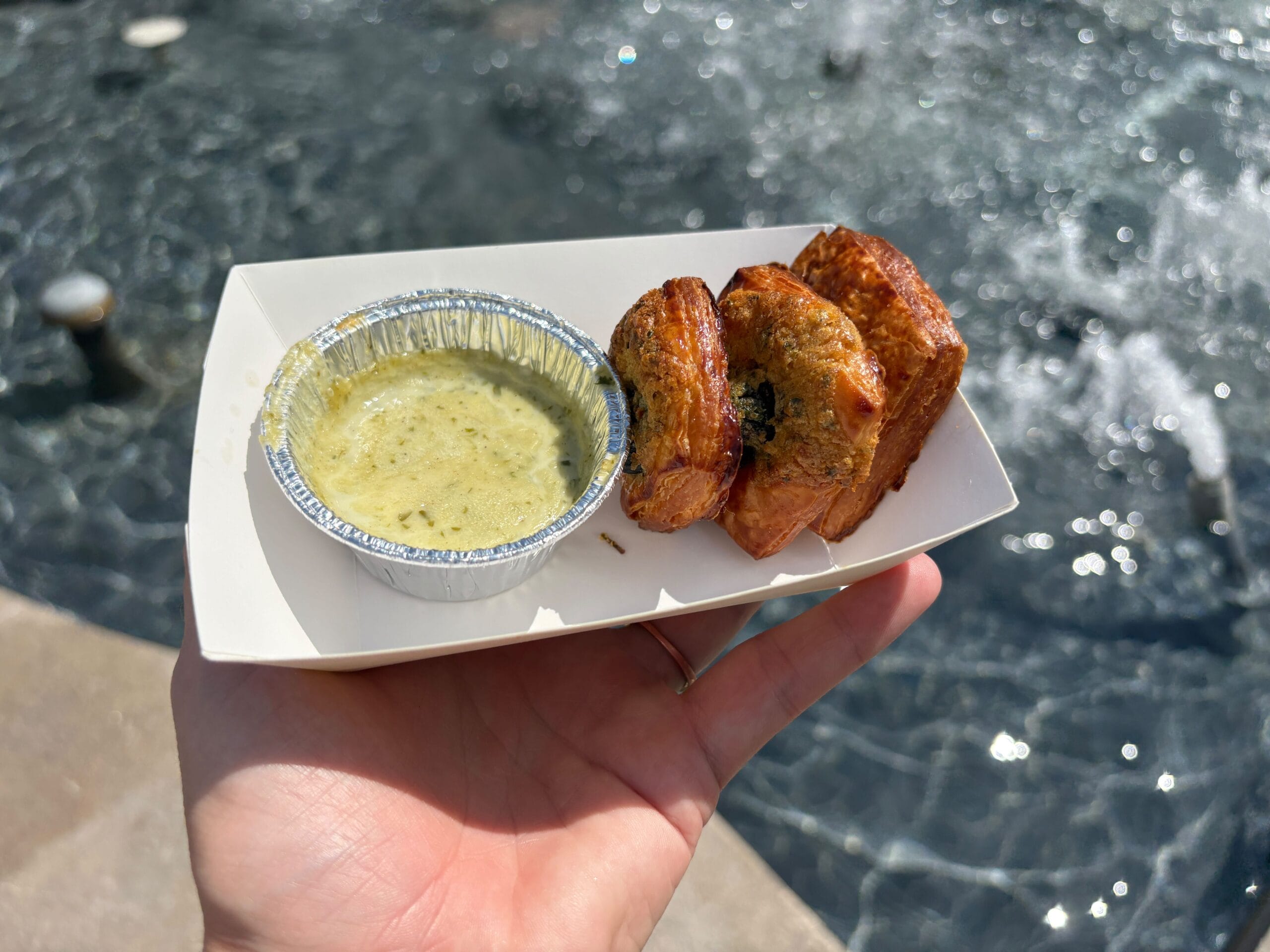 A hand holds fried plantains and New Food & Wine green sauce in front of a sunlit water background.