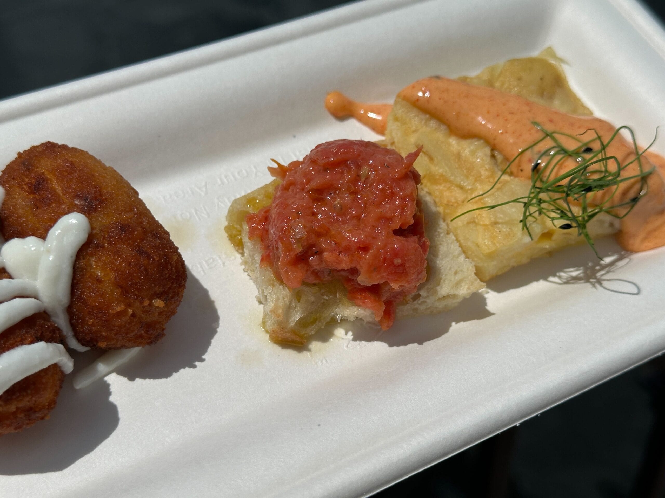 A paper plate with three Food & Wine appetizers: breaded bite with white sauce, tomato bread, and dill-topped orange sauce bread.