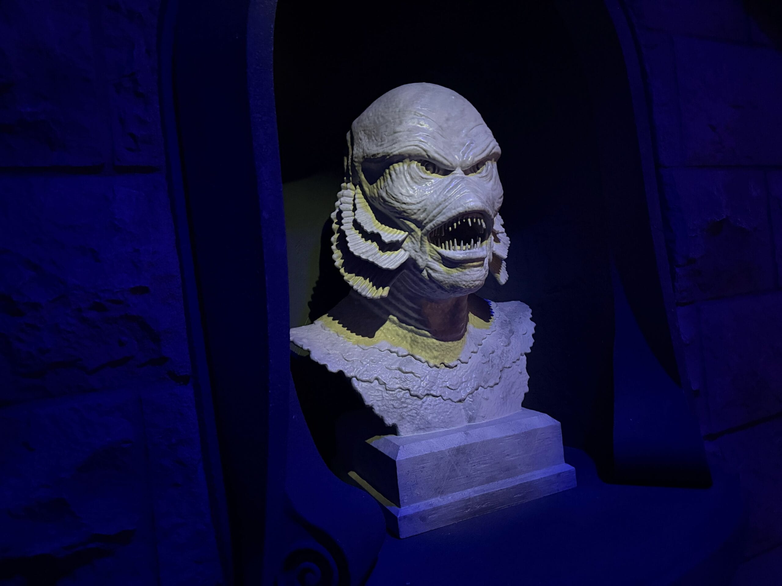 Bust of a creature with scales, sharp teeth, and gills evokes classic Universal Monsters in a dark, blue-lit alcove.