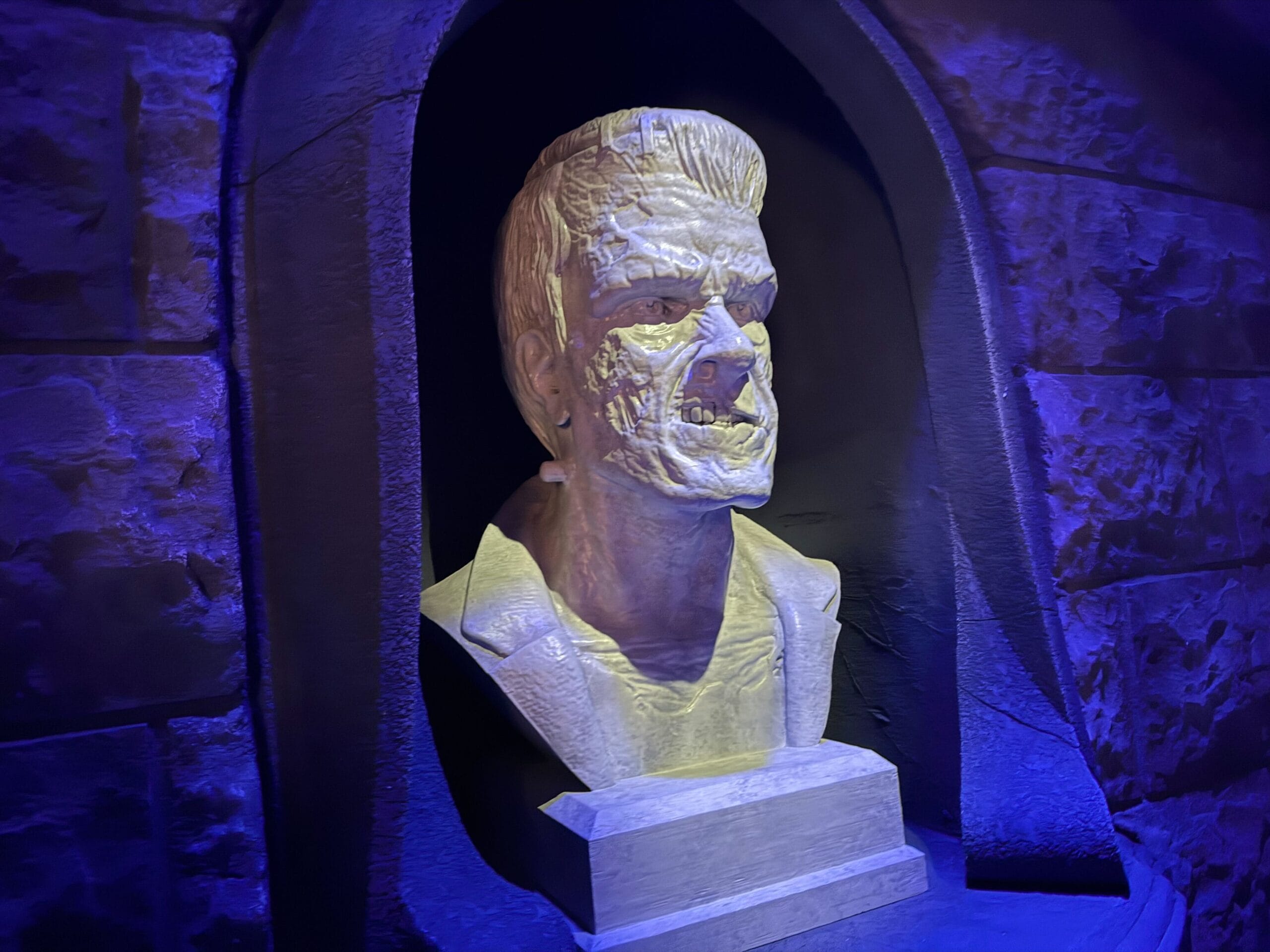 A stone bust of Frankenstein’s monster, icon of Universal Monsters, lit with blue and purple light in a stone alcove.