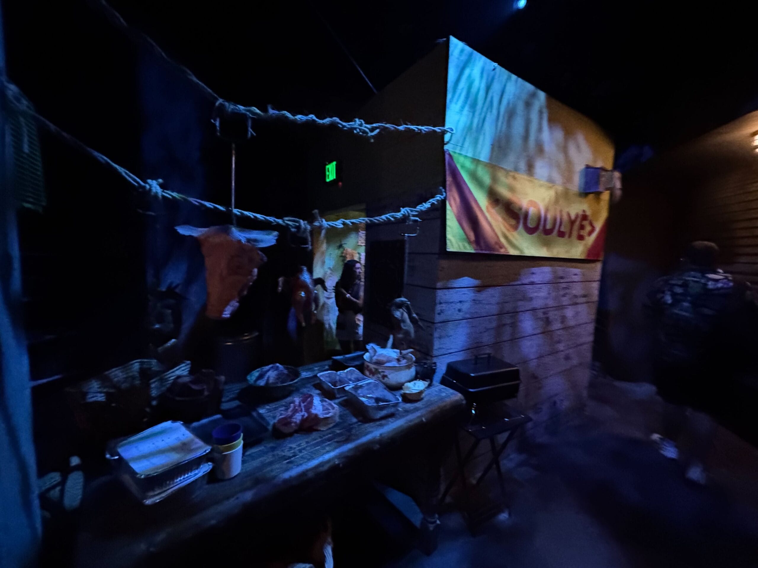 Dimly lit room with a table of food, hanging meat, and a yellow "SOULY'S" sign in the Exorcist Believer House.