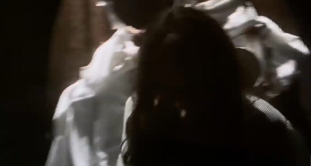 A person with long dark hair sits as another in white, like an Exorcist Believer House scene, appears behind them.