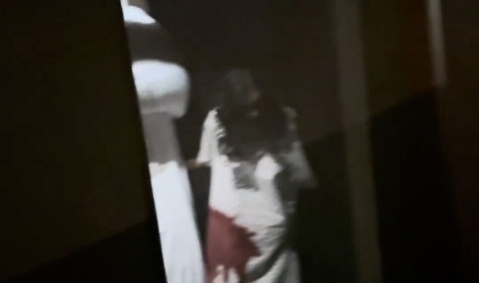 A person in a white dress with a large red stain stands in a hallway during a Universal Horror Unleashed house tour.