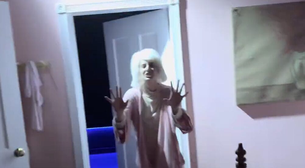 A person in light pajamas stands in the Exorcist Believer House doorway at night, hands raised in surprise or alarm.