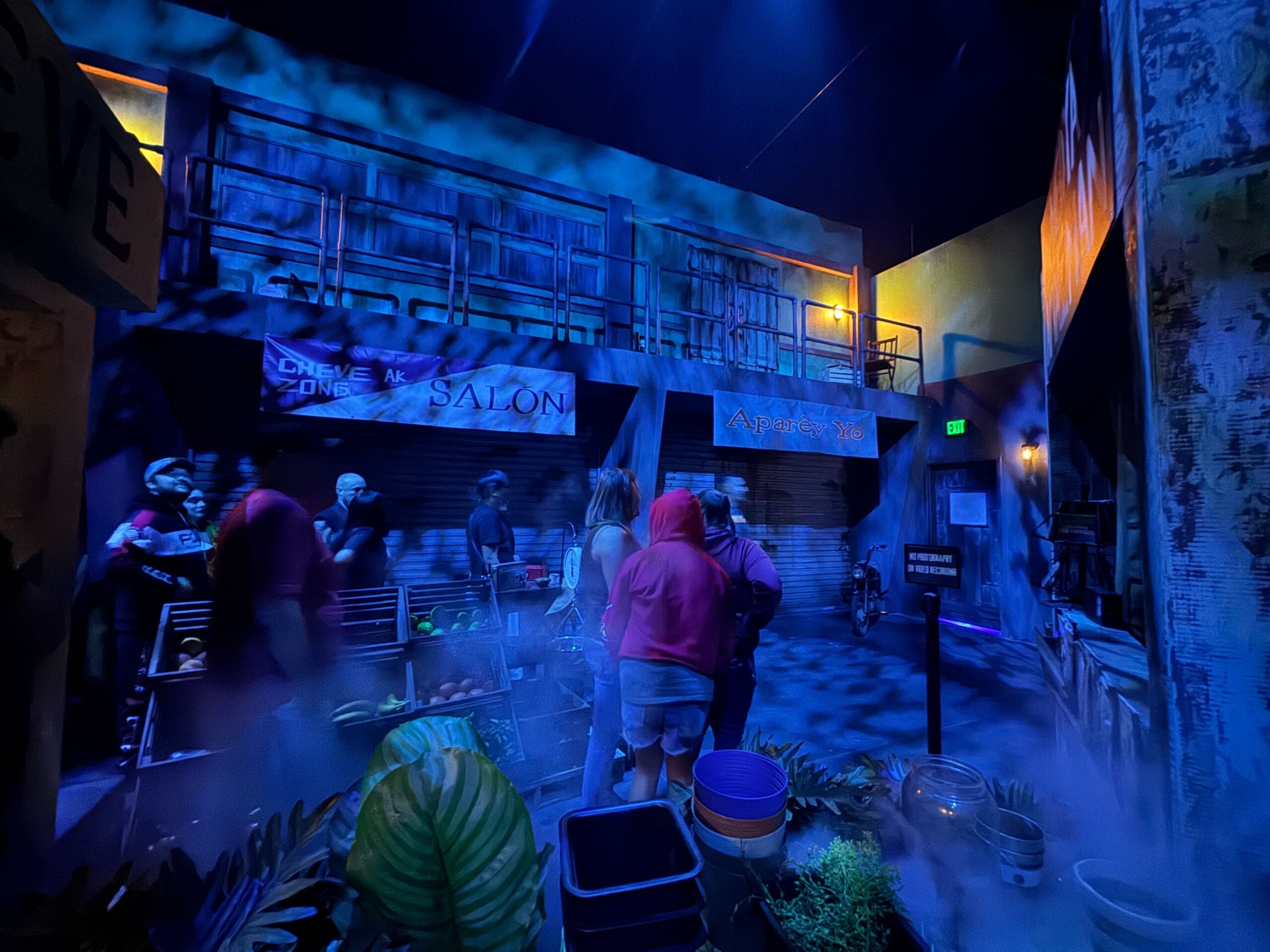 People gather in a dimly lit, foggy, blue-toned urban set on the Universal Horror Unleashed full tour.