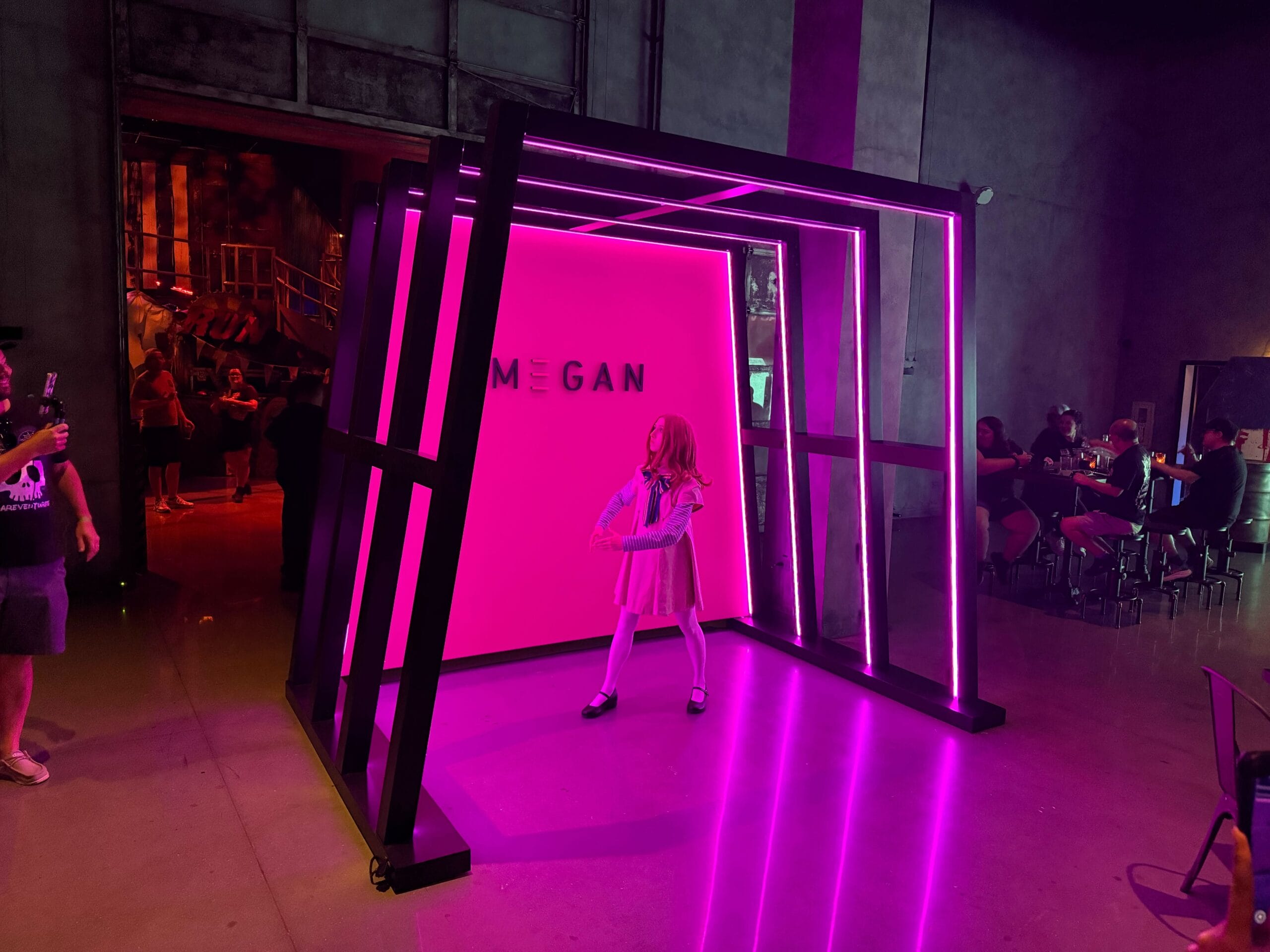 A person in a MEGAN costume poses in a neon-lit pink booth at Universal Horror Unleashed, with onlookers nearby.