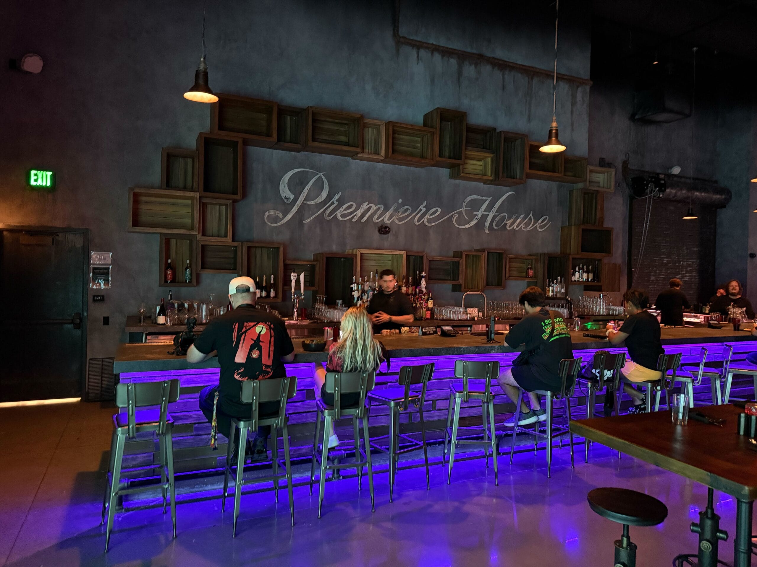 People sit at a bar with a purple-lit counter, browsing the Premier House menu in a modern, dimly lit space.