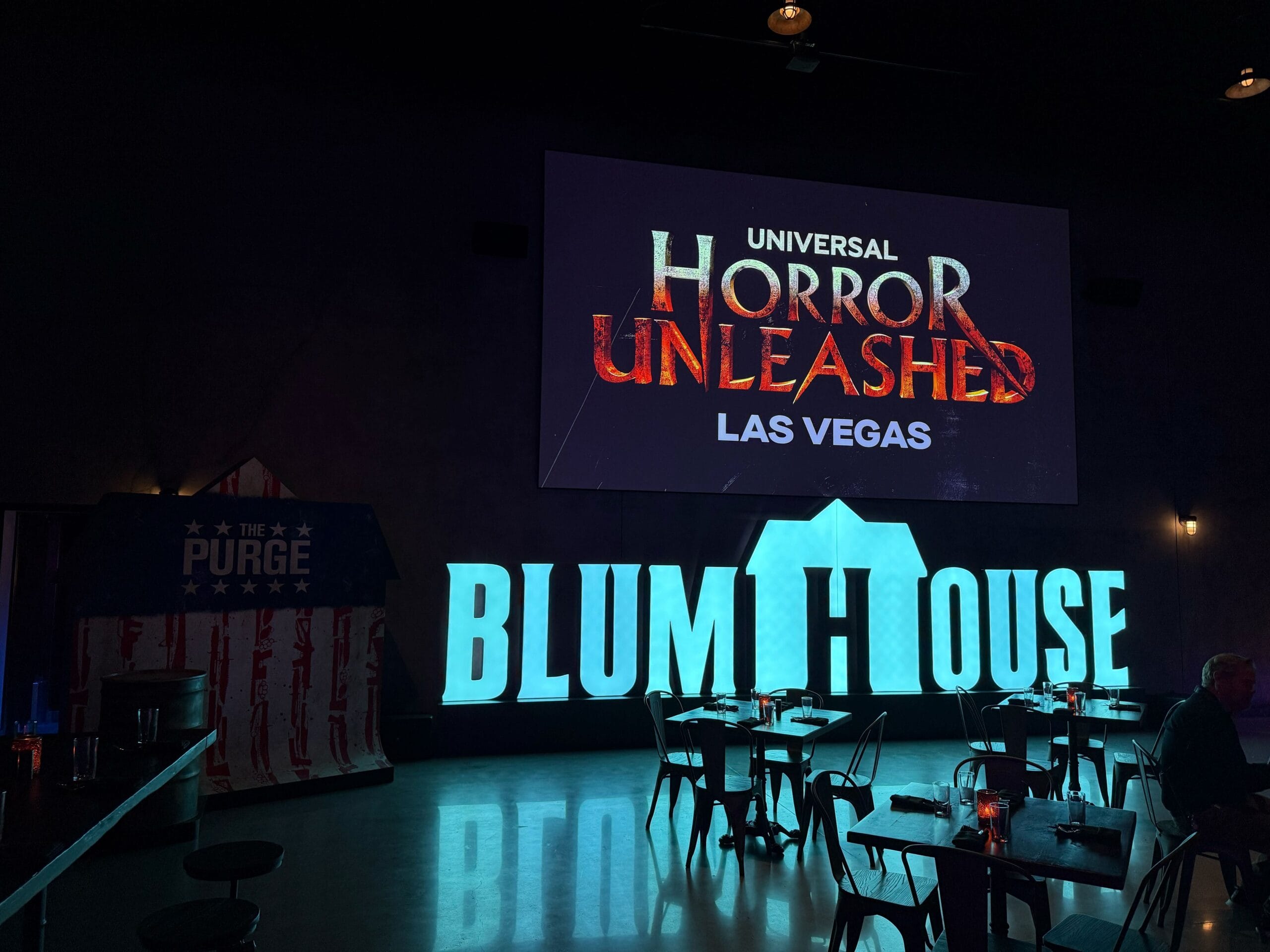Dimly lit room with "Blumhouse" lights and Universal Horror Unleashed Las Vegas menu prices projected on screen.