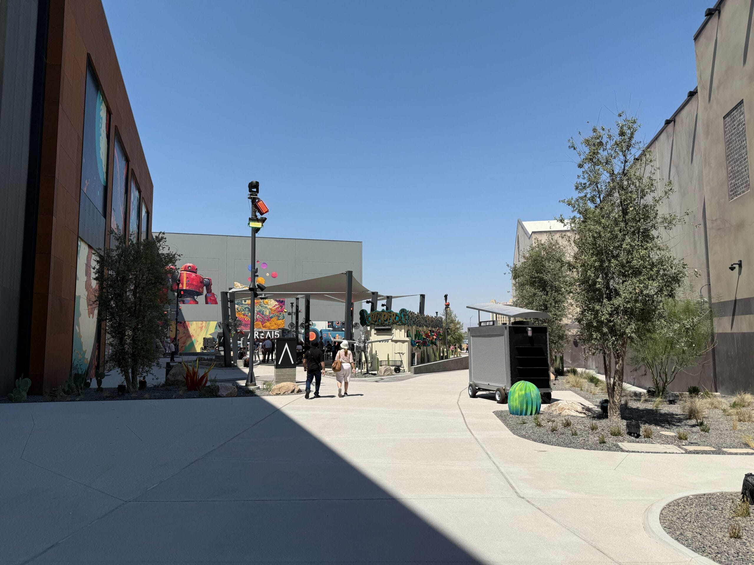 People stroll a sunny Las Vegas pathway lined with murals, plants, and modern buildings under a clear blue sky.