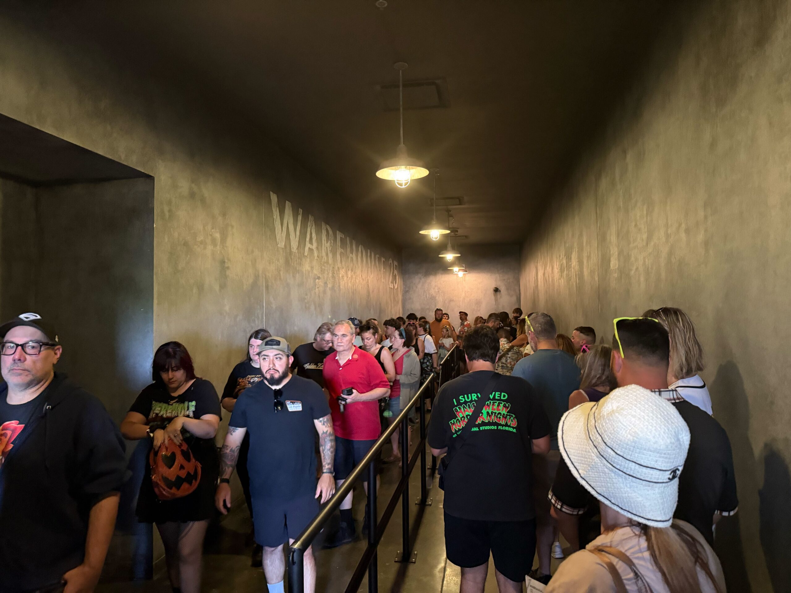 A crowd waits in a dimly lit hallway for the Universal Horror Unleashed Full Tour, standing in line with black railings.