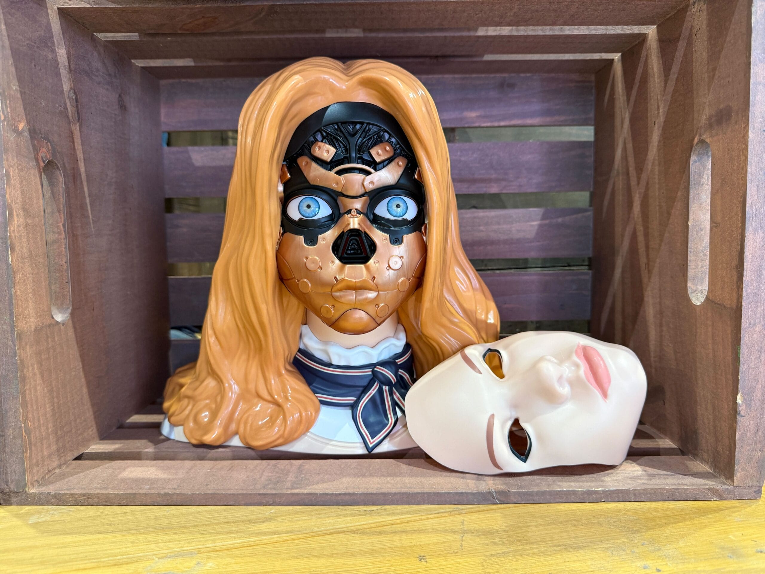 A robotic female M3GAN head with its synthetic skin removed, displayed inside a wooden crate.