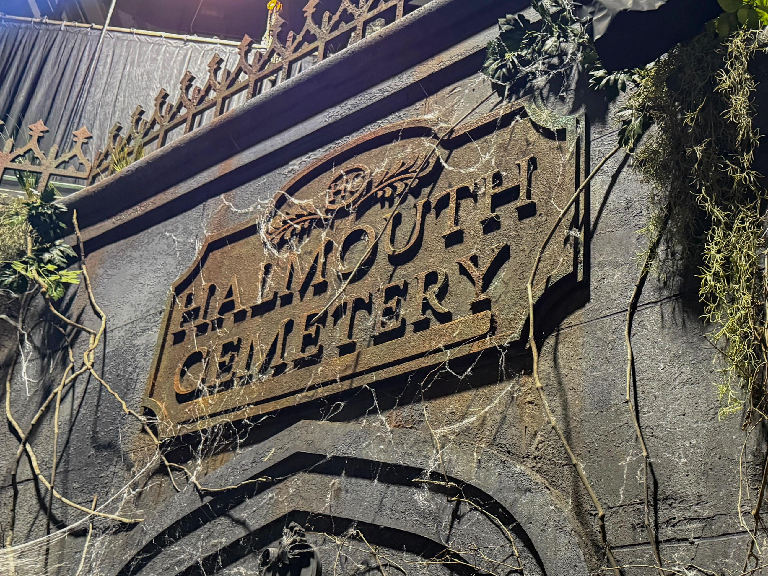 A rusty sign reading "FALMOUTH CEMETERY" hangs on a wall overrun by vines—HHN Houses would envy its eerie decay.