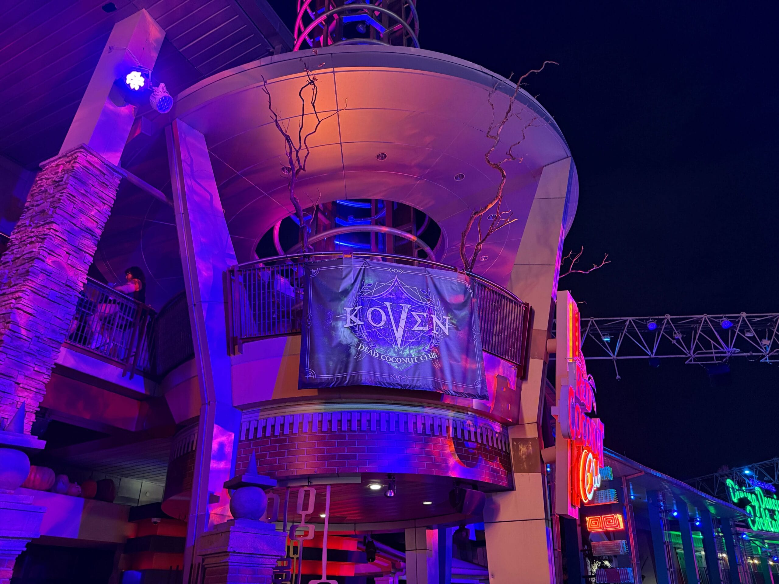 A balcony at night displays a “Koven” sign with purple lighting and branches at the Dead Coconut Club entertainment venue.