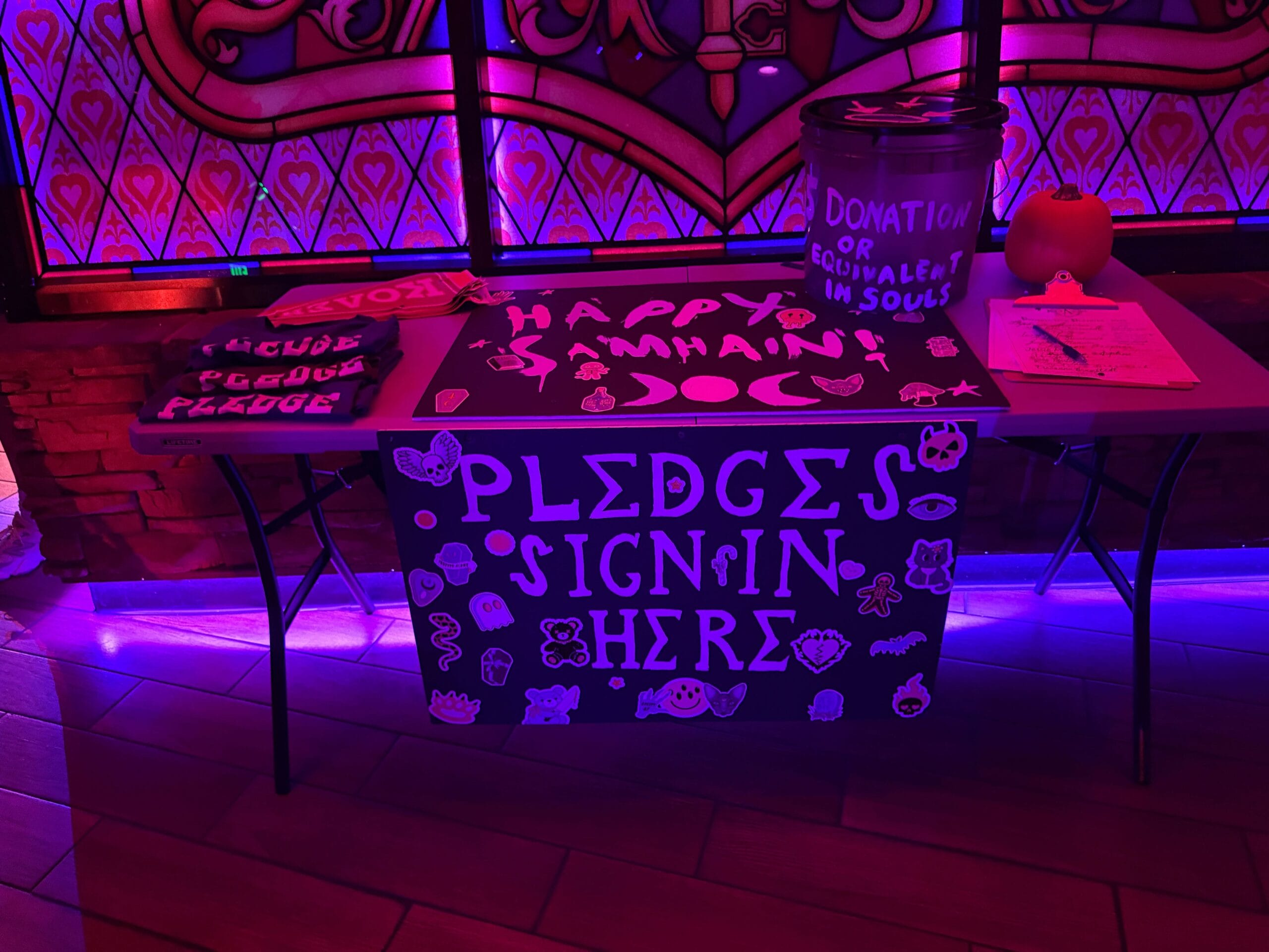 A table with Sorority Witches’ "Pledges Sign In Here" sign, donation bucket, and "Happy Samhain" decor under purple lights.