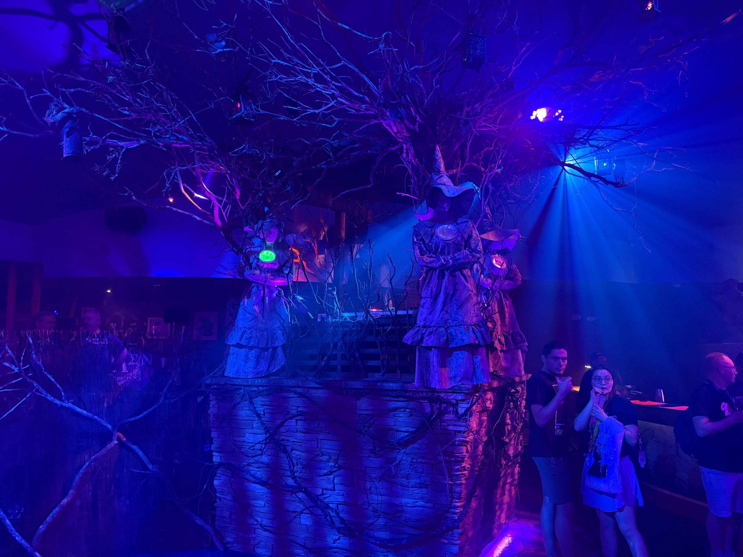 Two people stand near eerie tree-like creatures at the Dead Coconut Club, bathed in blue and purple light on the Sorority Witches tour.