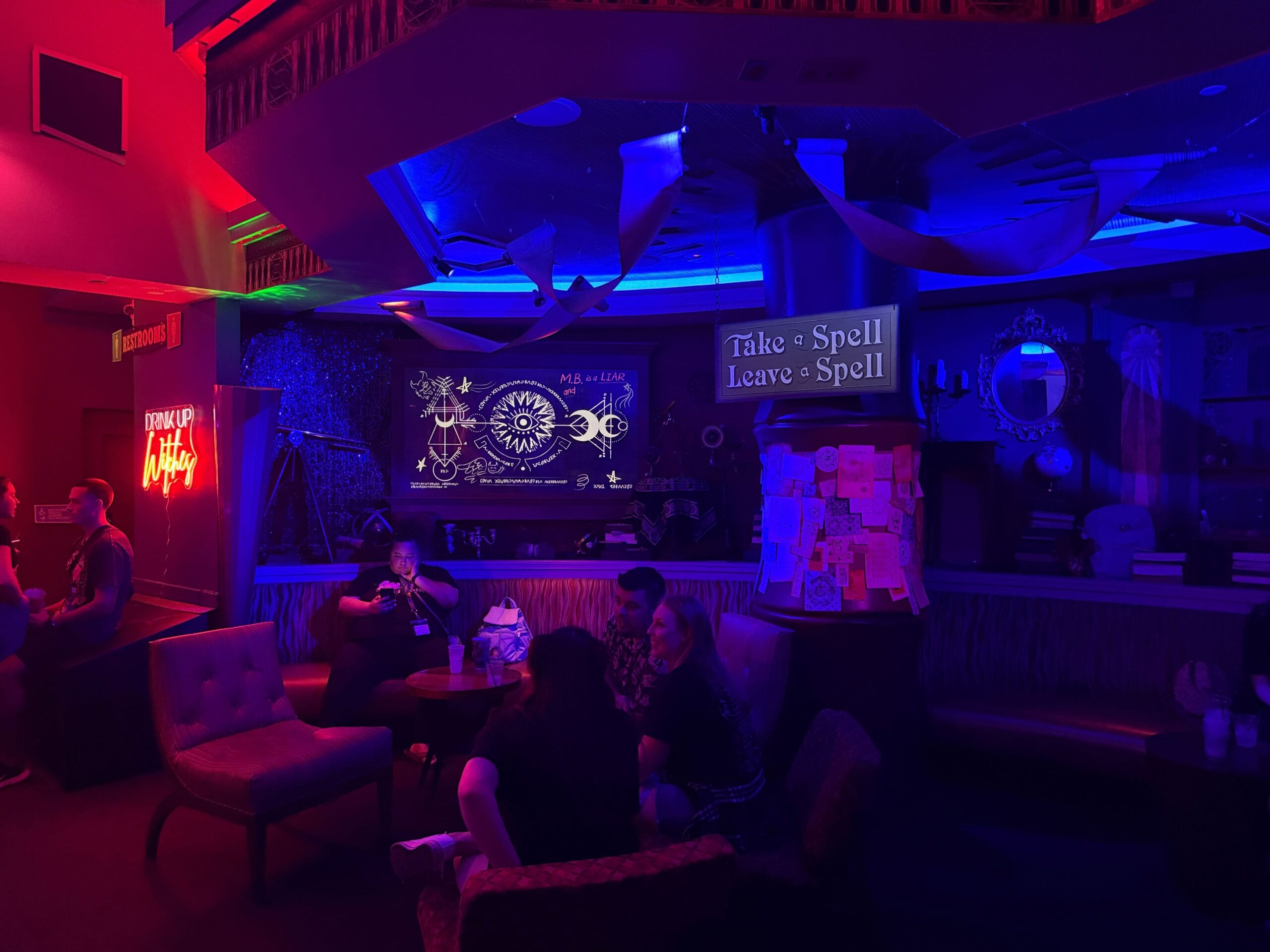 Dimly lit lounge with neon lights, mystical decor, and Sorority Witches vibe—sign reads “Take a Spell, Leave a Spell.”