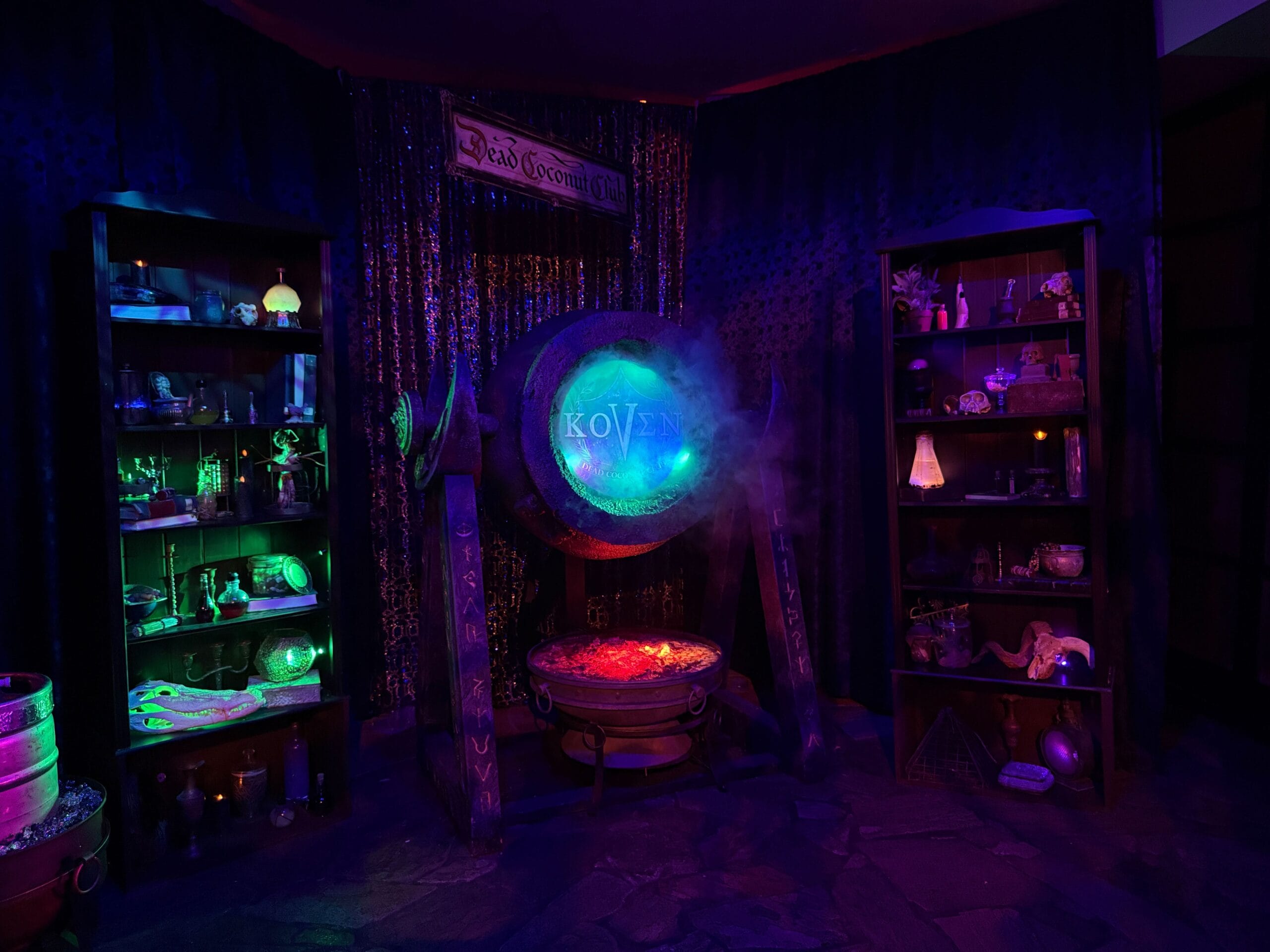 A mystical, dimly lit room at Dead Coconut Club with sorority witches, magical items, and a glowing cauldron by a large gong.