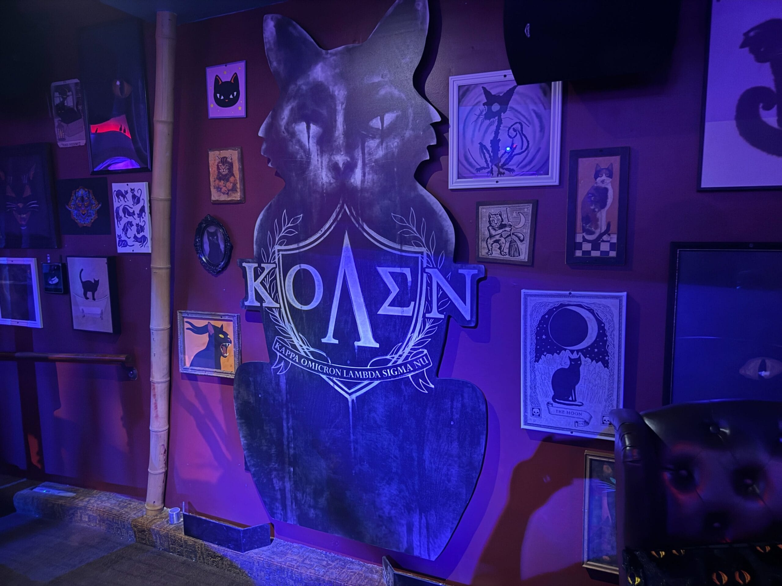 A wall decorated with black cat art, framed pictures, and a large Sorority Witches sign with Greek letters ΚΟΛΣΝ.