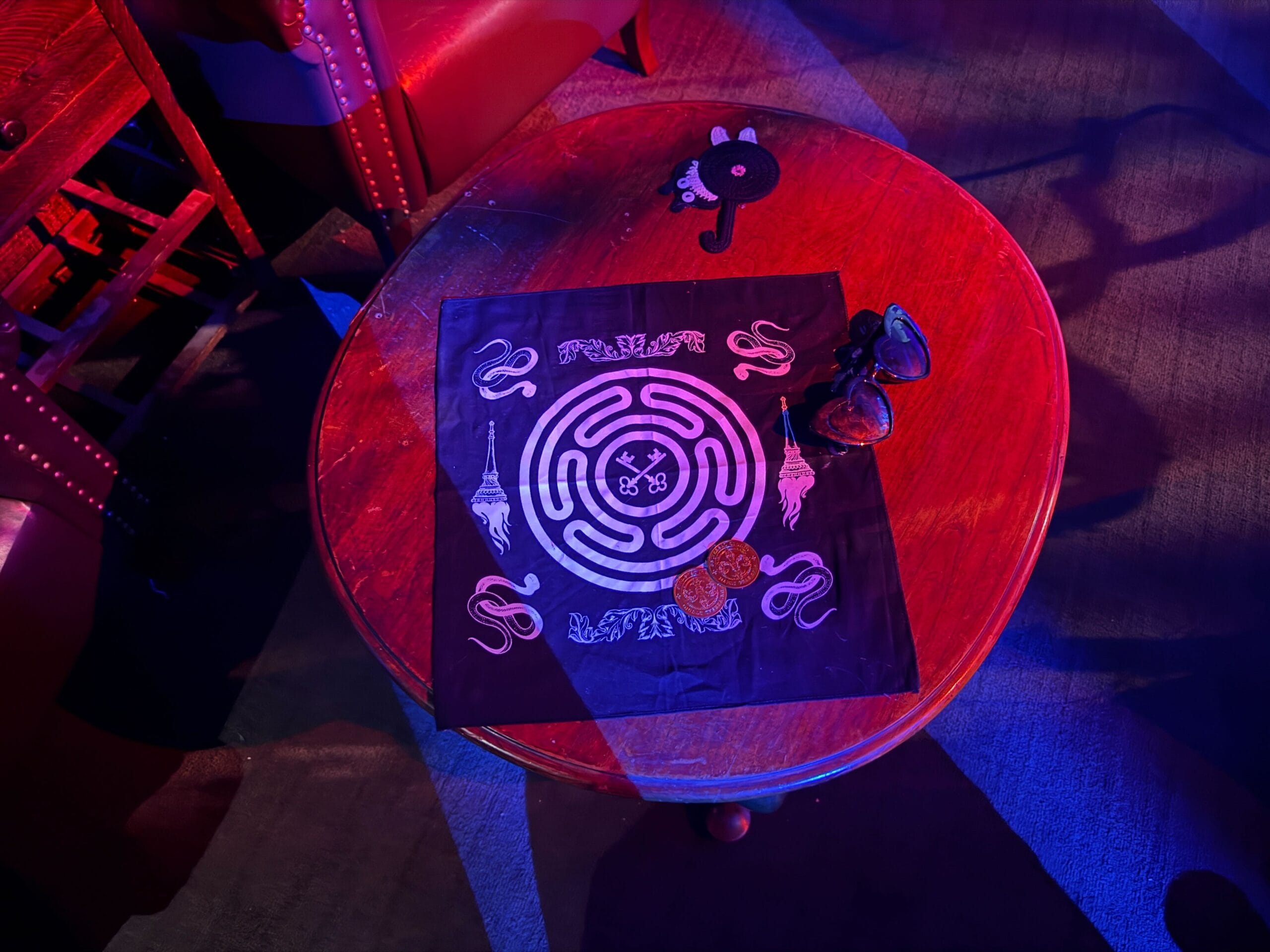 A round wooden table with a maze-patterned cloth, sunglasses, a coin, and a Sorority Witches plush bat keychain.