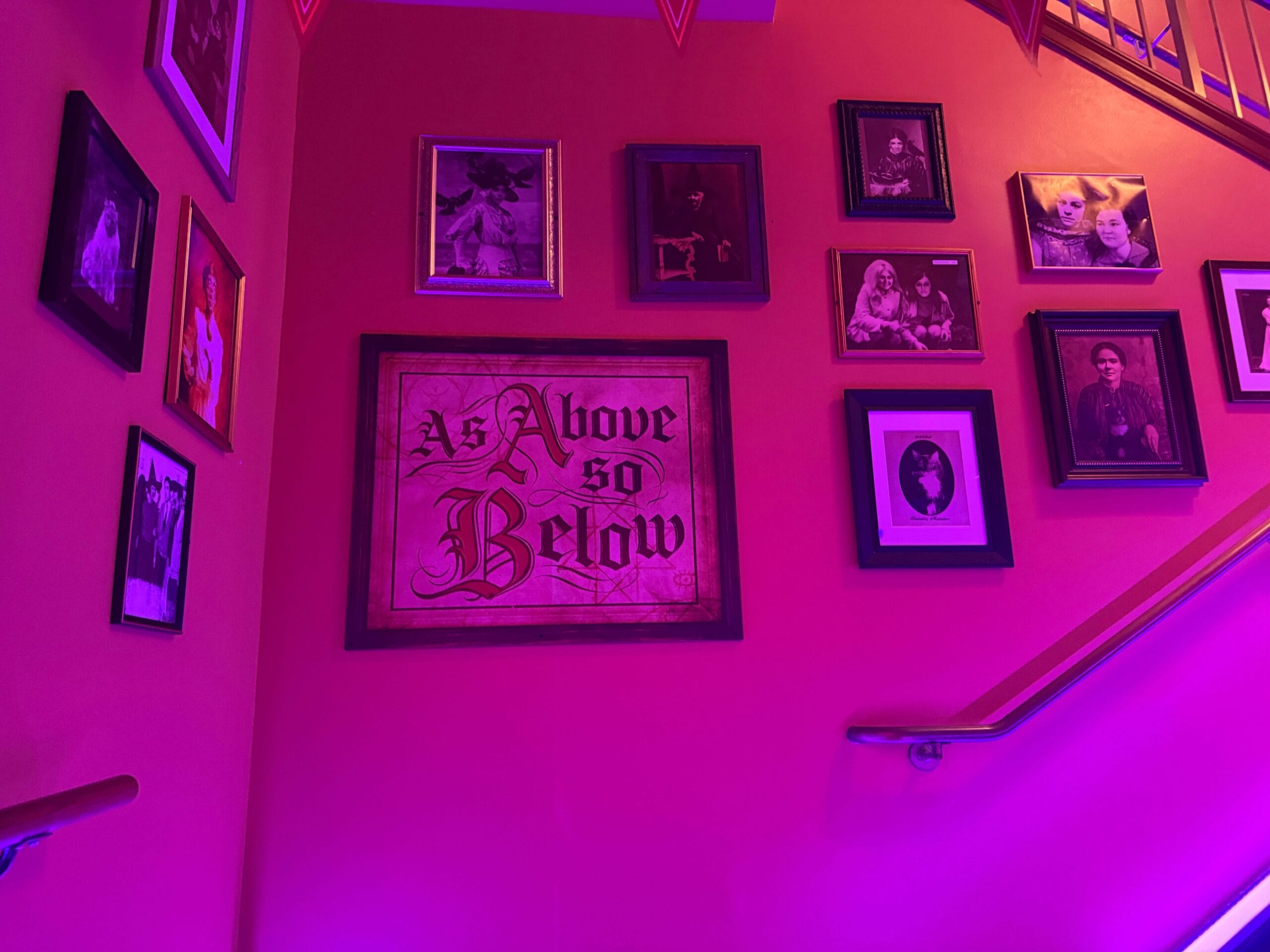 A staircase wall with framed photos and a large "As Above So Below" sign glows under purple Dead Coconut Club lighting.
