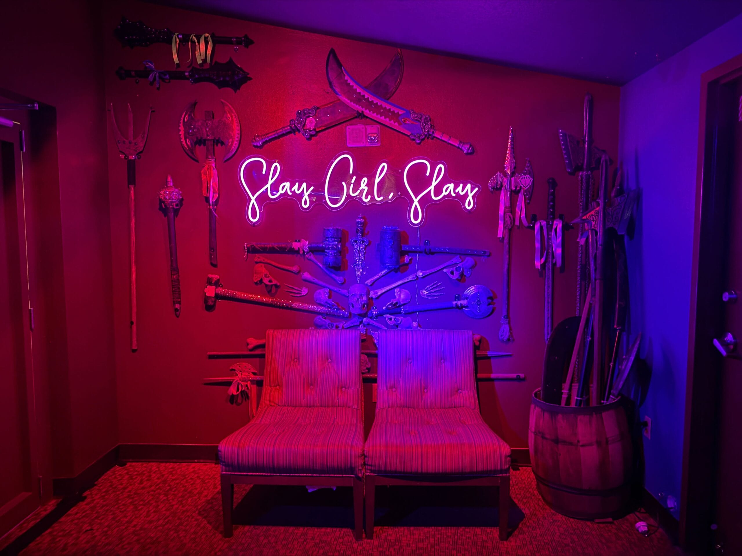 Purple-lit room with medieval weapons, neon “Slay Girl Slay” sign, and two chairs—fit for Sorority Witches.
