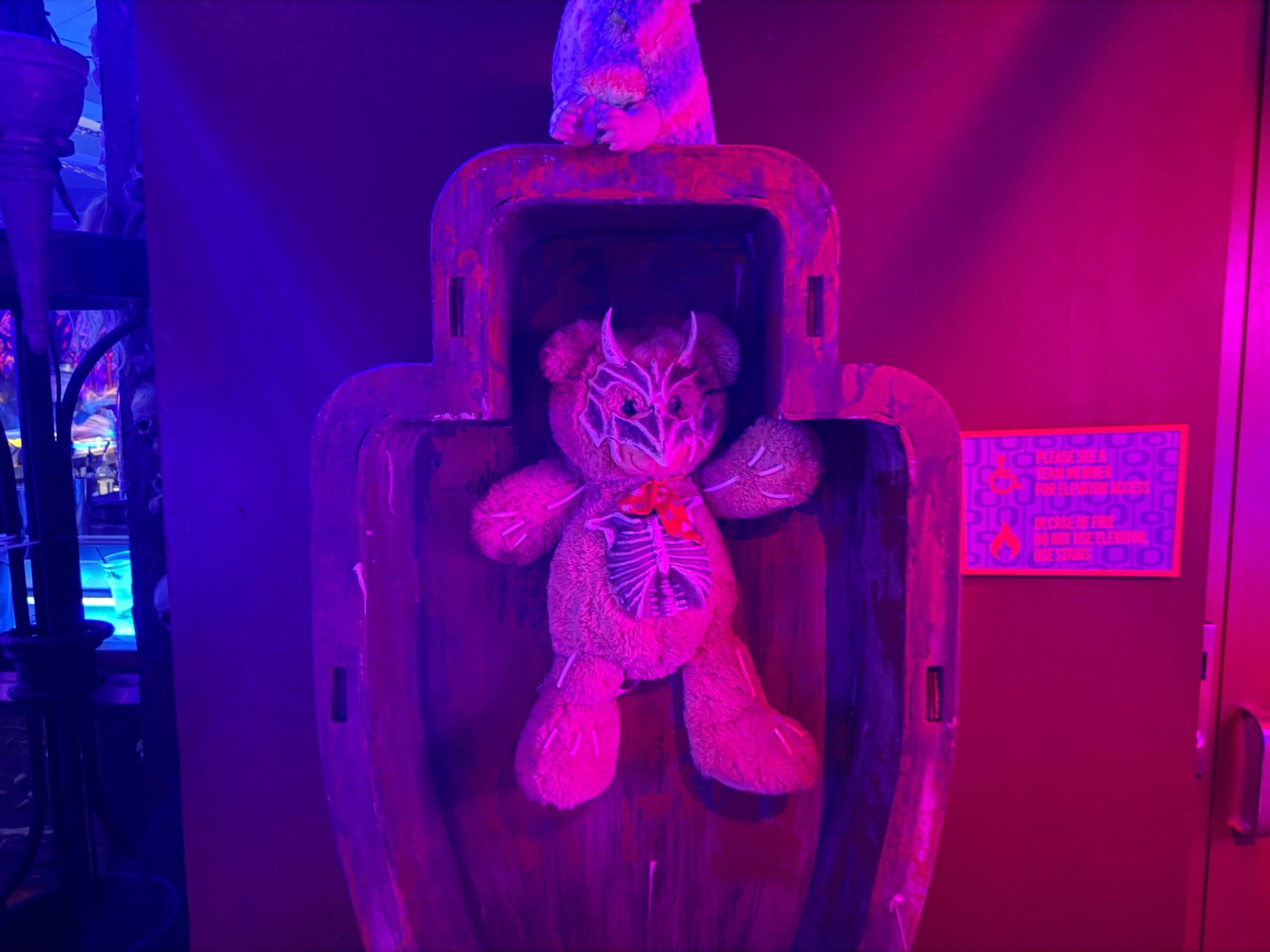 A teddy bear with a skeletal design and horns is displayed in a coffin-like case at the Dead Coconut Club under purple lighting.