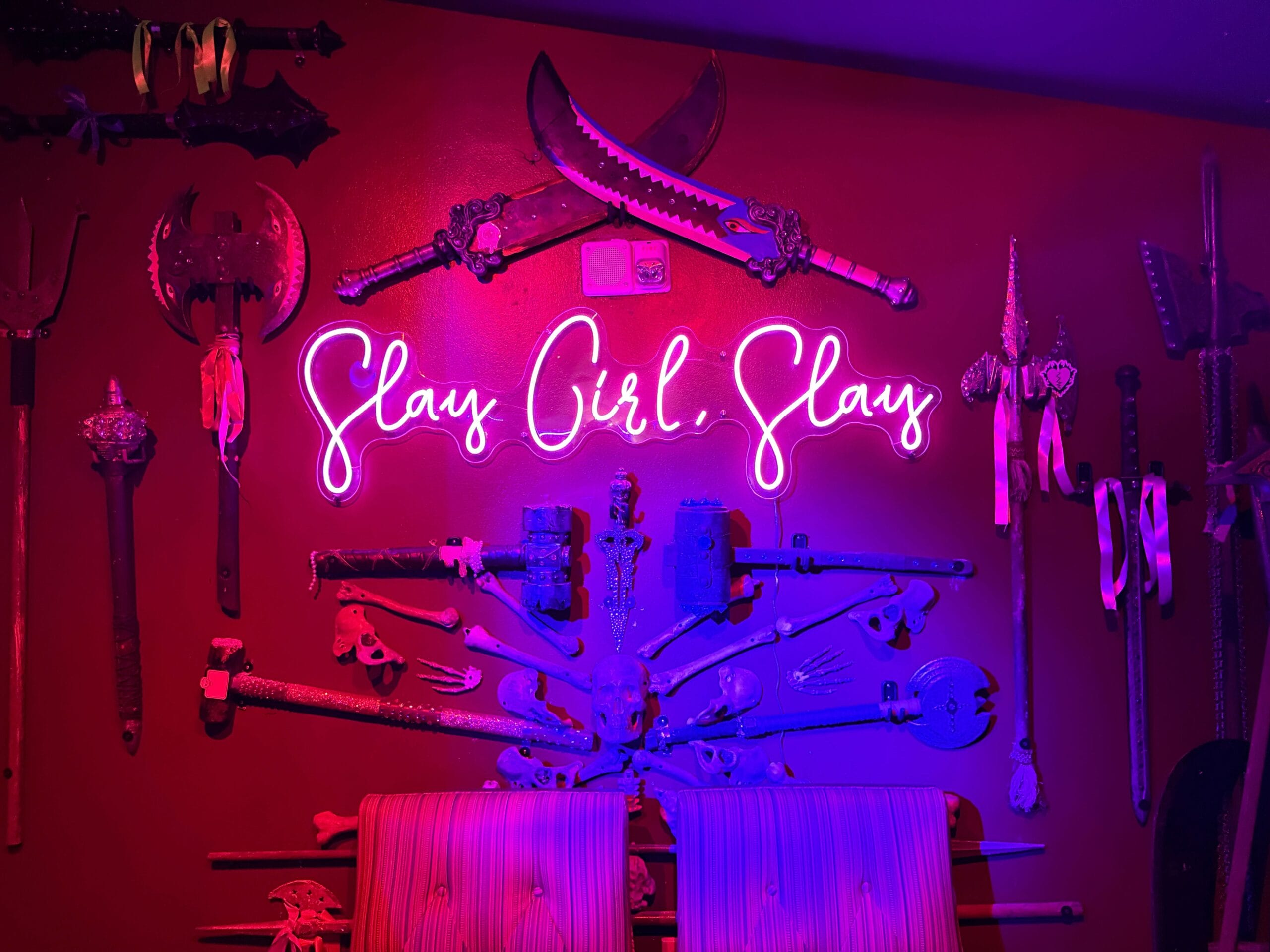 Neon "Slay Girl Slay" sign glows on a red wall, echoing Sorority Witches energy amid medieval weapons.
