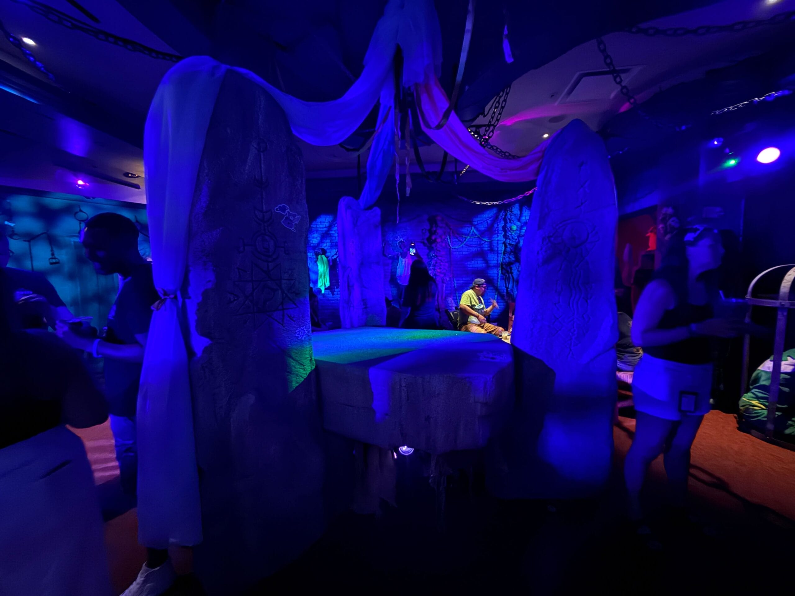 Dimly lit room with stone pillars, draped cloth, and Sorority Witches from the Dead Coconut Club mingling throughout.