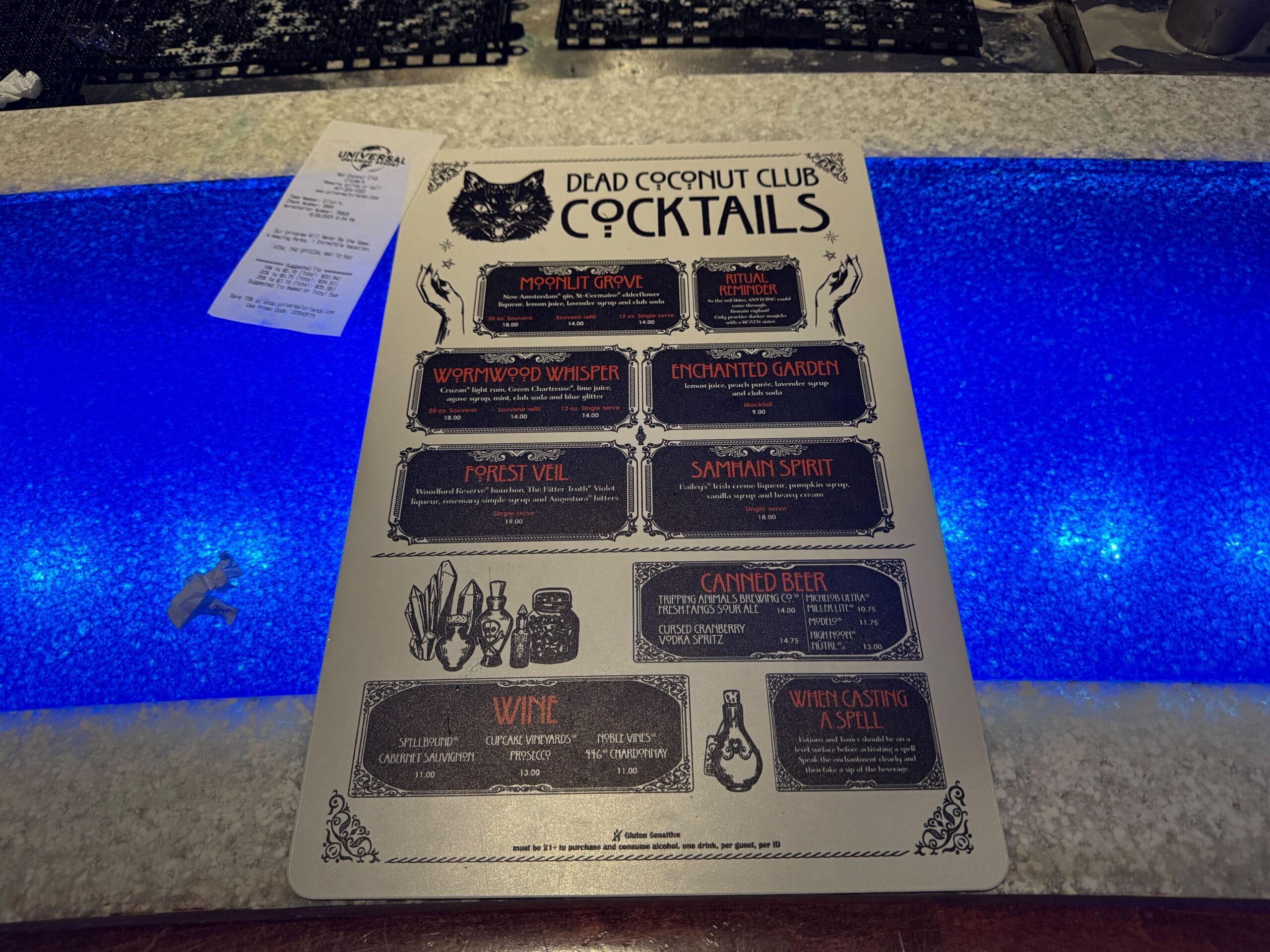 A Dead Coconut Club cocktail menu on a glowing blue bar, featuring Sorority Witches-themed drink options.