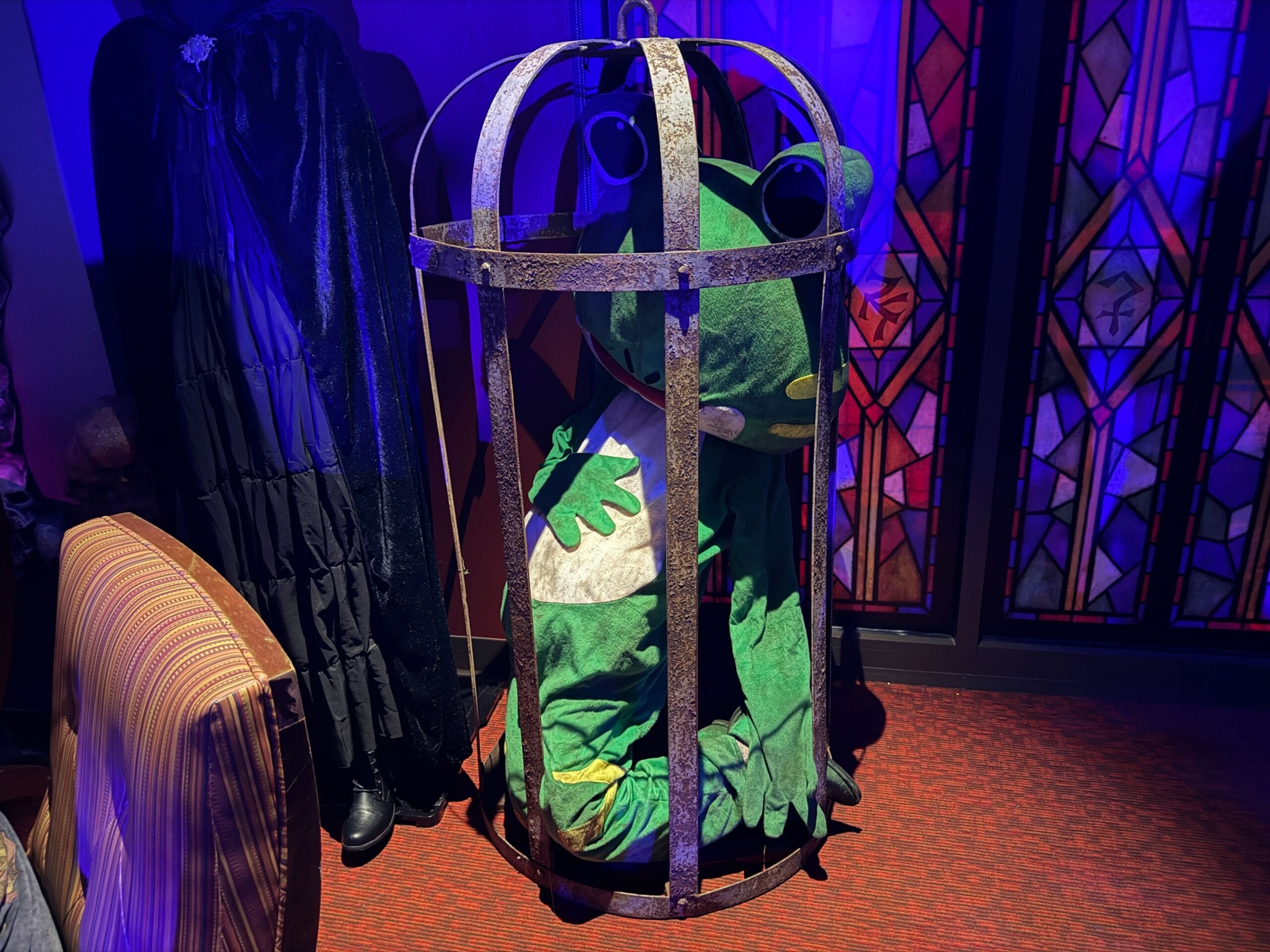A person in a green frog costume sits caged in a dim room at Dead Coconut Club, stained glass panels glowing.