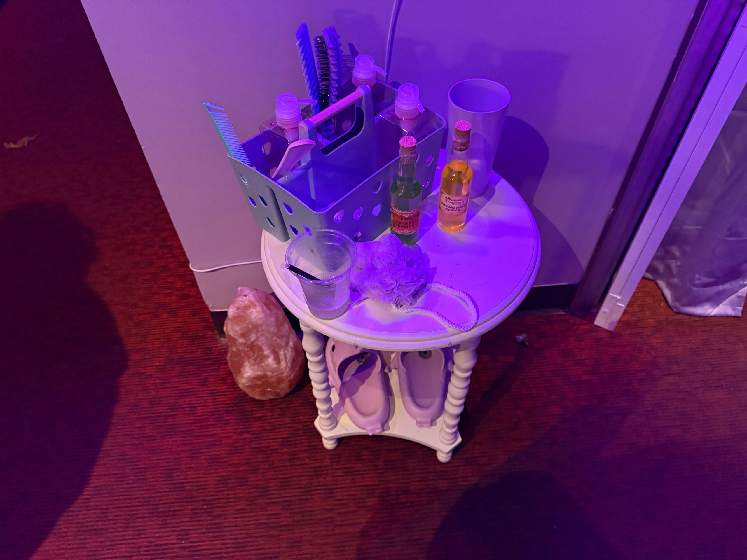 A small round table at the Dead Coconut Club holds drinks, a cup, toiletries, a loofah, and a lamp on a red carpet.