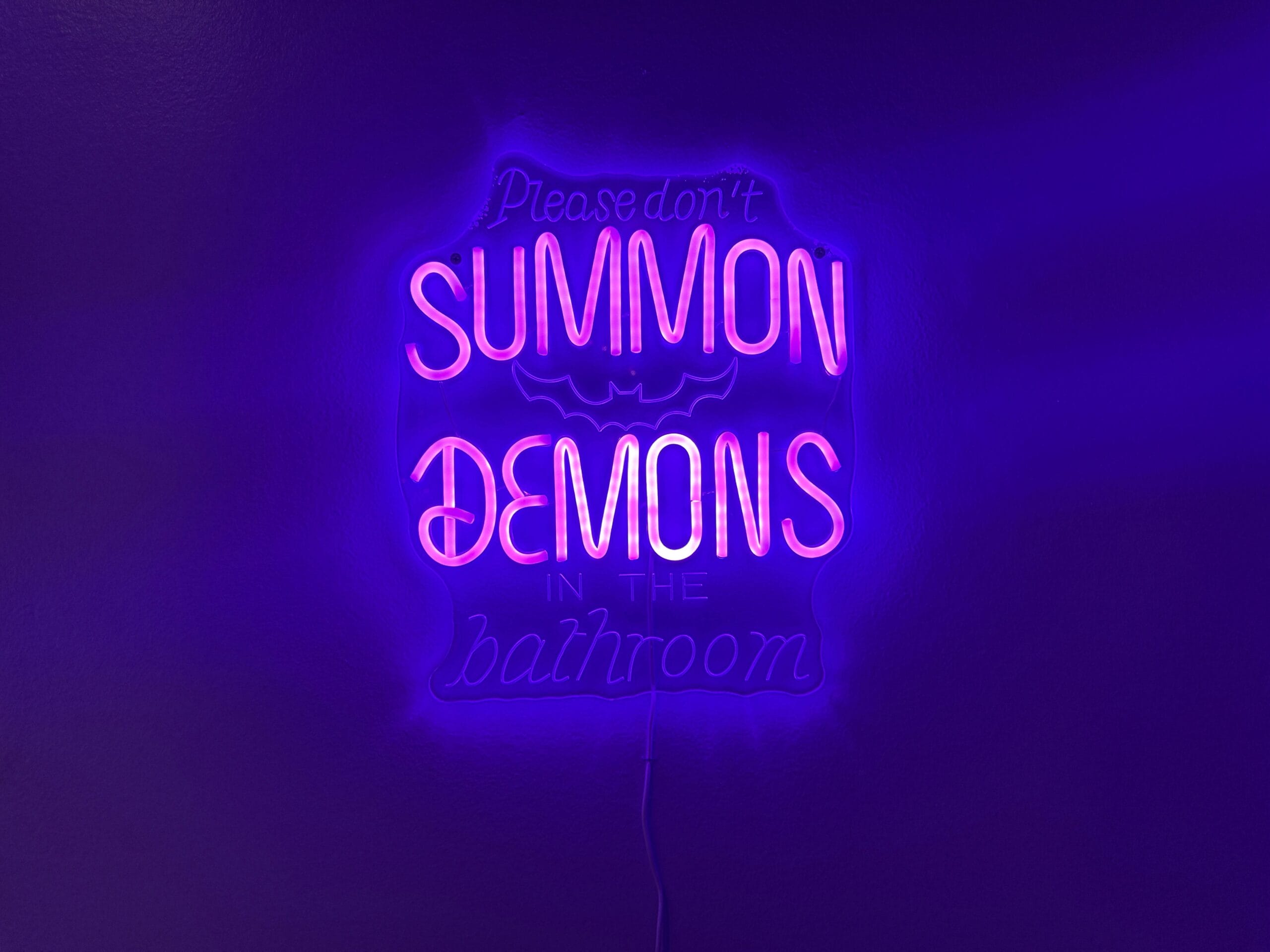 Neon sign at Dead Coconut Club reads "Please don't summon demons in the bathroom" in pink and purple light.